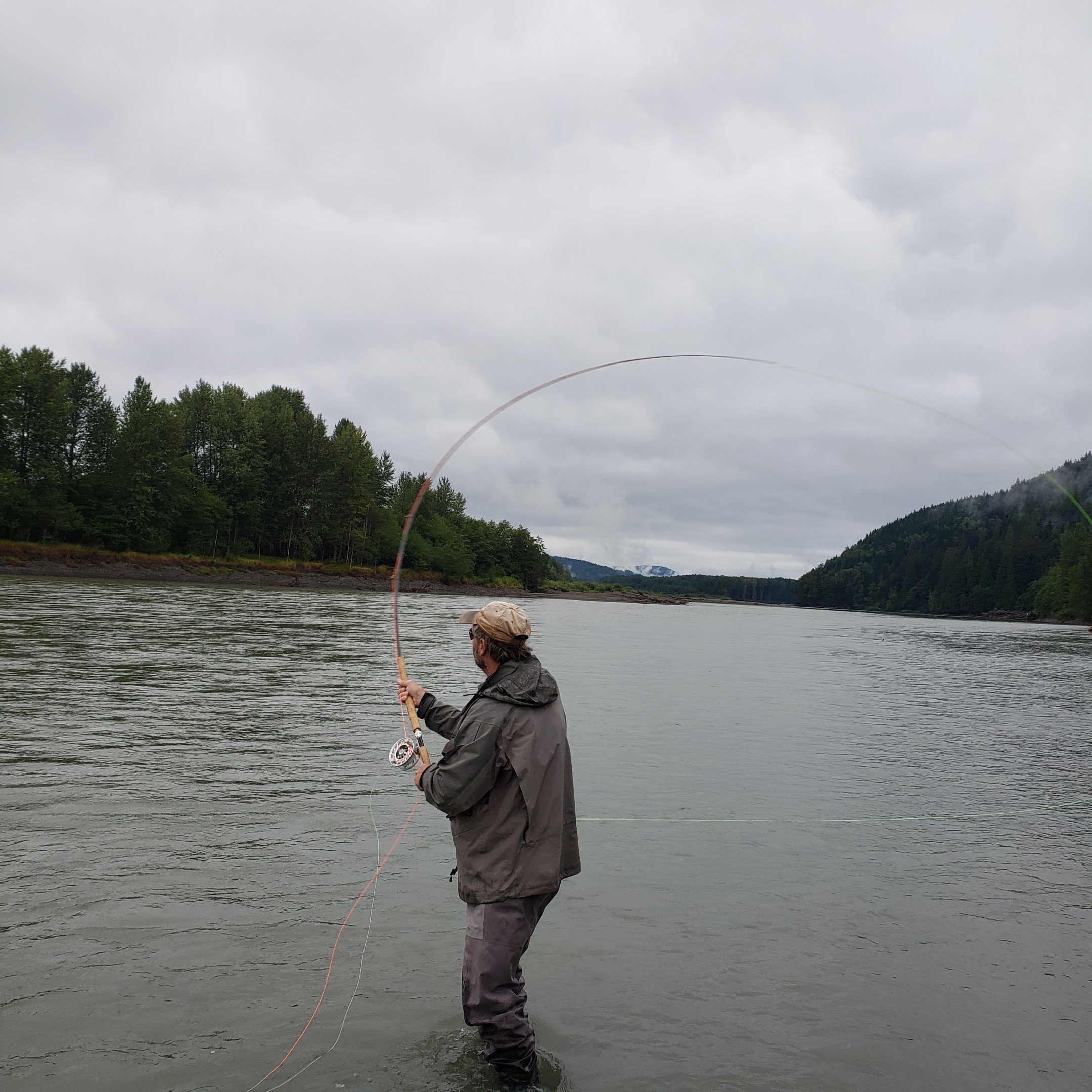 A person standing in a river, fishing with a cloudy sky above, and trees and hills in the background.