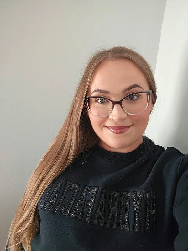 A young woman with long straight hair wearing glasses and a black t-shirt with text is taking a selfie indoors in front of a plain light-colored wall.