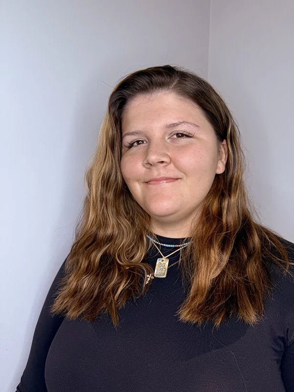 A woman with wavy, shoulder-length brown hair, wearing a black top and layered necklaces, smiling with one eye slightly closed, against a plain light-colored wall background.