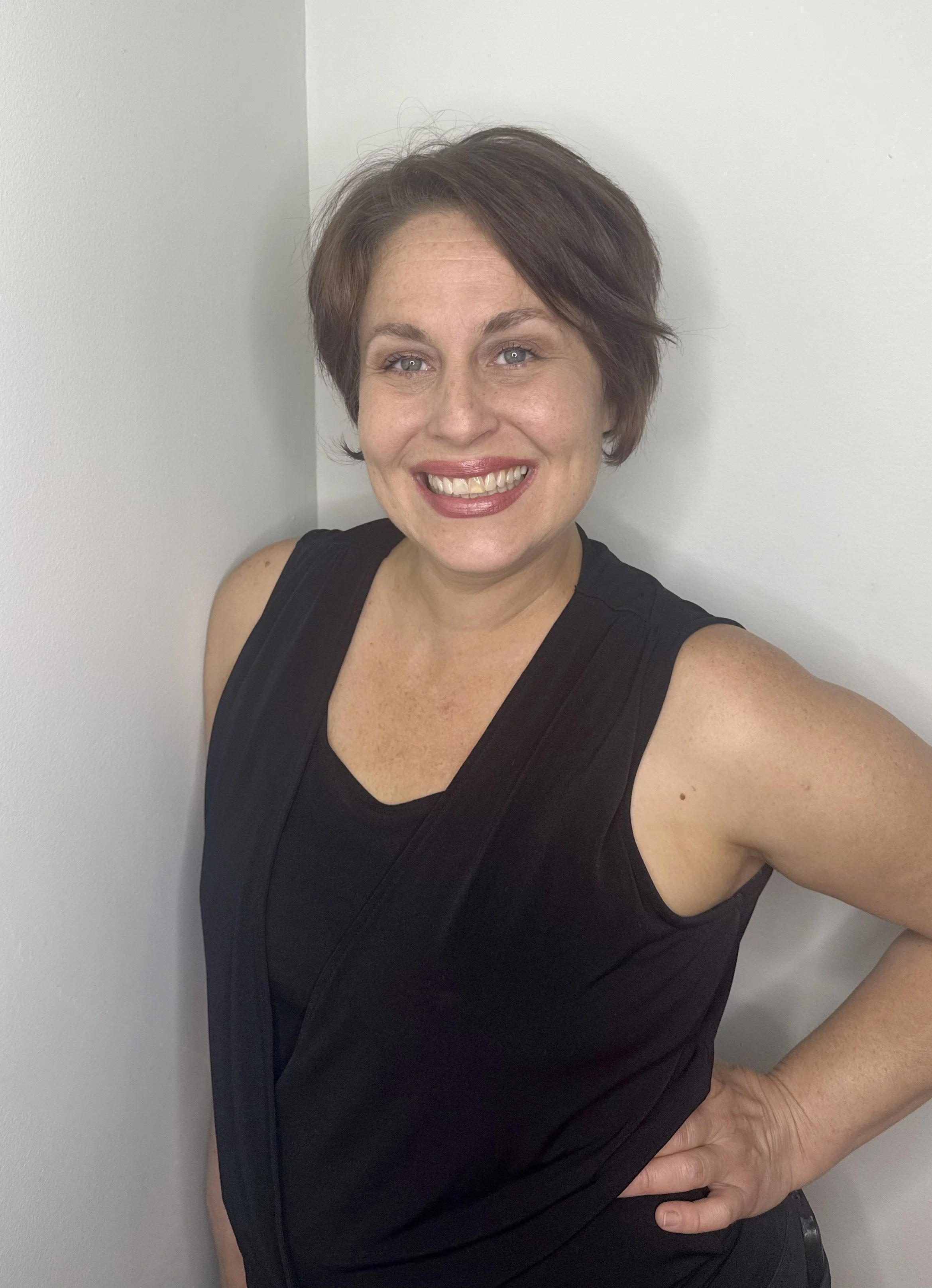 A woman with short brown hair, smiling, standing with one hand on her hip, wearing a sleeveless black top, against a white wall.