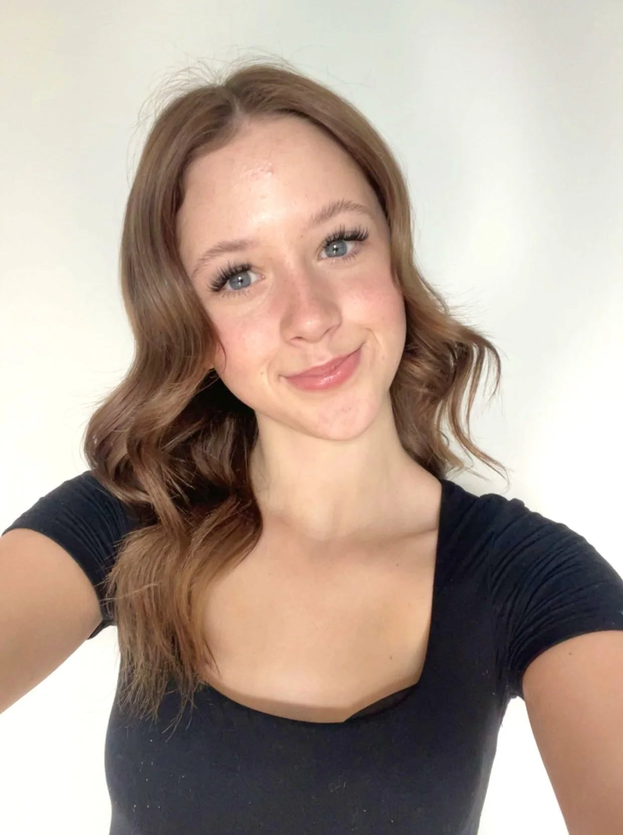 A young woman with wavy, shoulder-length auburn hair and light blue eyes, smiling slightly, wearing a black short-sleeve top, standing against a plain white background.