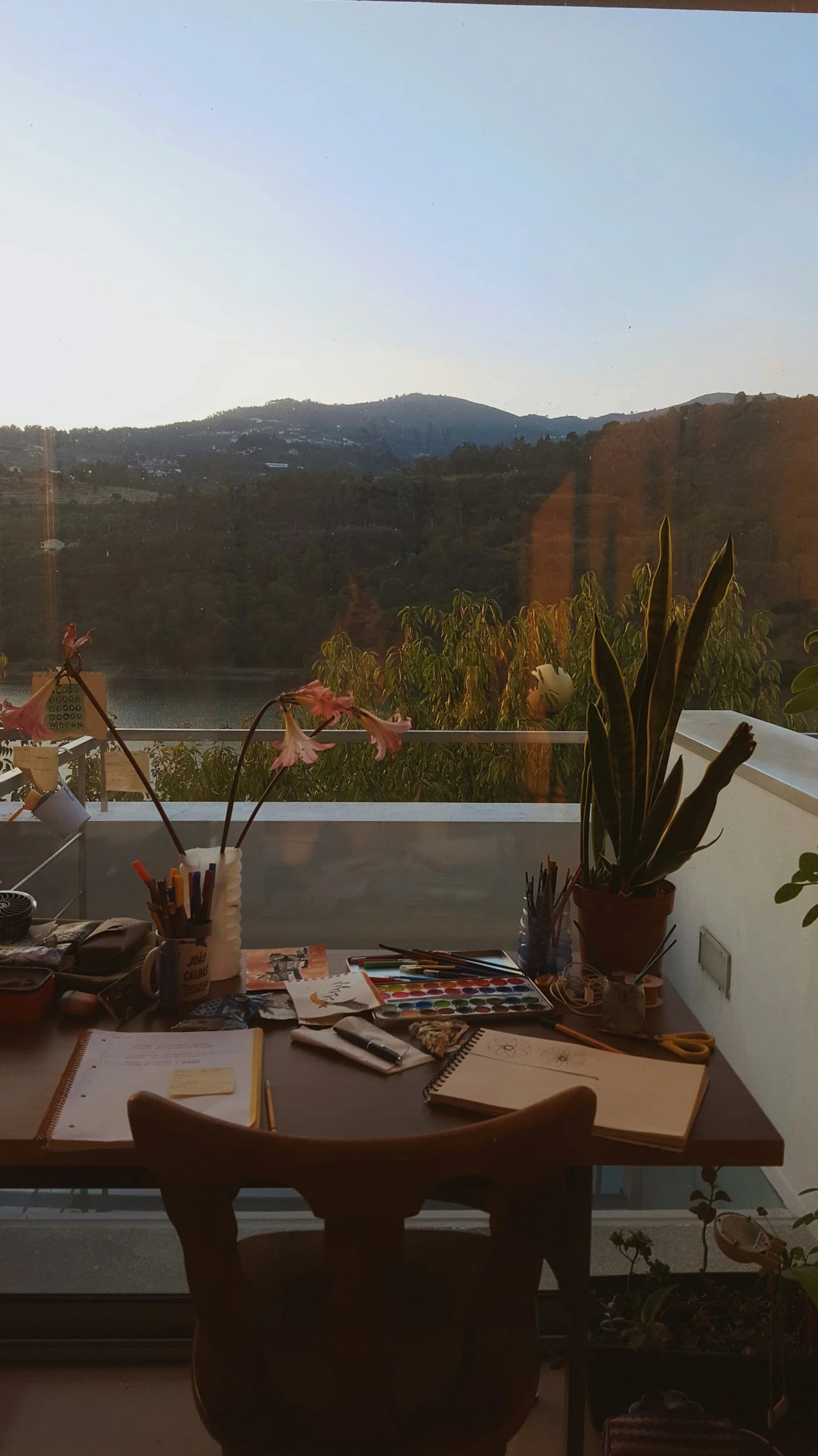 View of a room with a desk cluttered with art supplies, notebooks, scissors, and a mug, overlooking a balcony with plants and a scenic hillside landscape under a clear sky.
