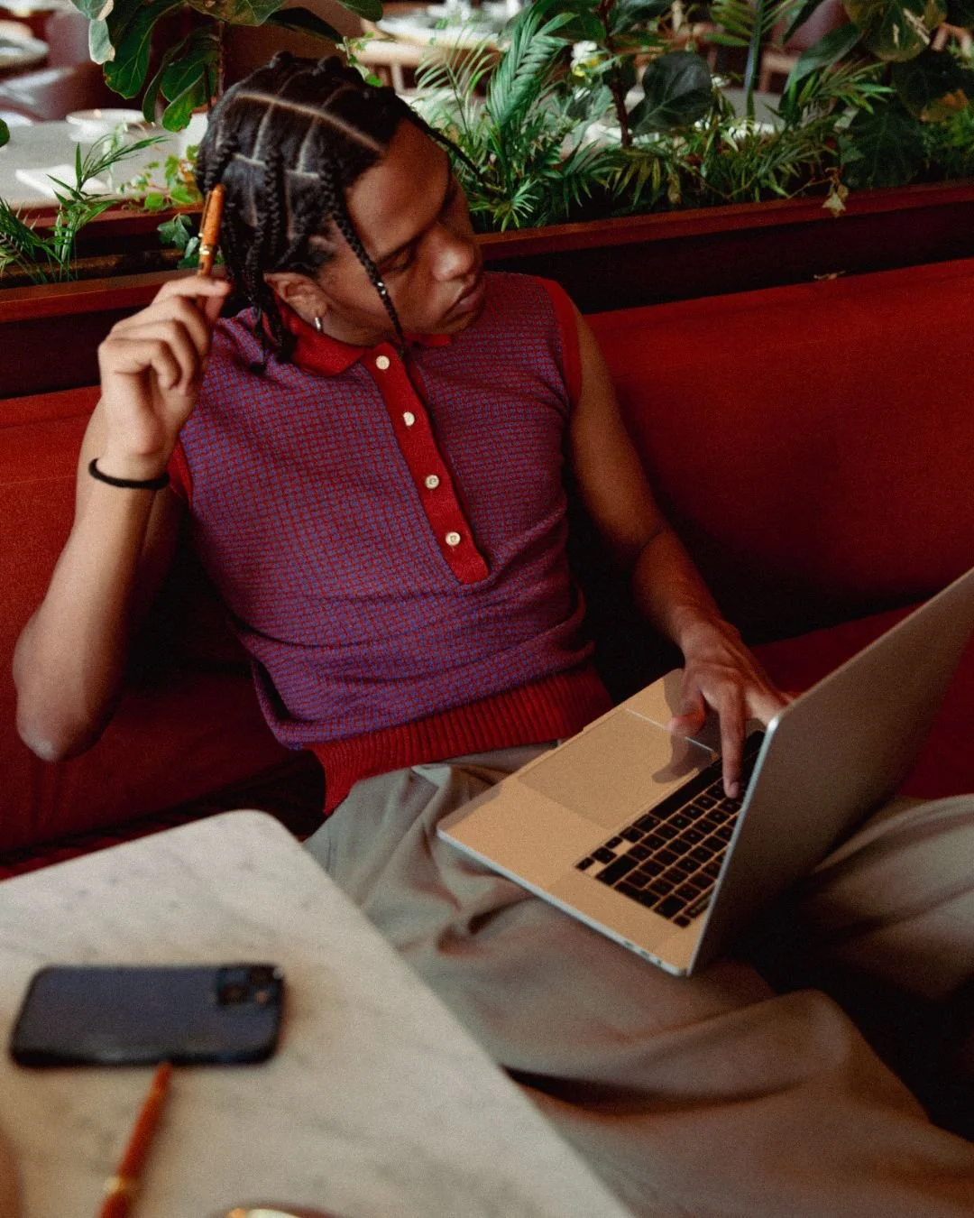 A young man with braids sitting on a red booth in a restaurant, using a silver laptop. He wears a sleeveless red and blue patterned shirt, beige pants, and has a black hair tie on his wrist. There is a smartphone and a pen on the table in front of him, and plants are visible behind him.