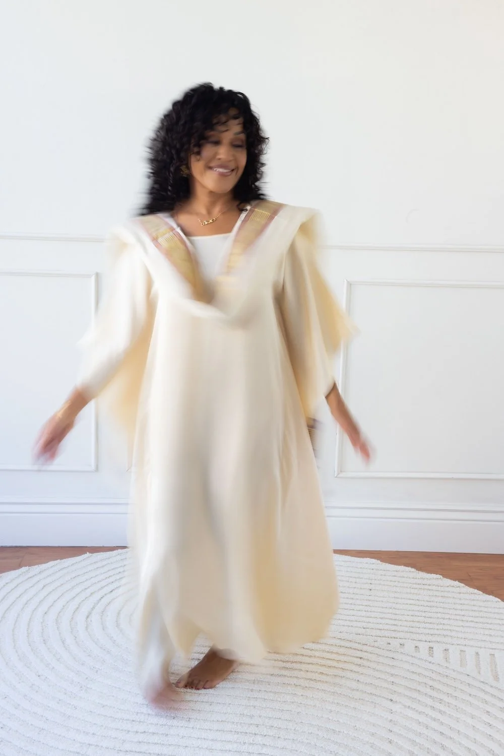 Maya Jaguar, an interfaith reverend dancing at Love Church in a cream-colored, elegant dress while barefoot on a white rug in a room with white walls.