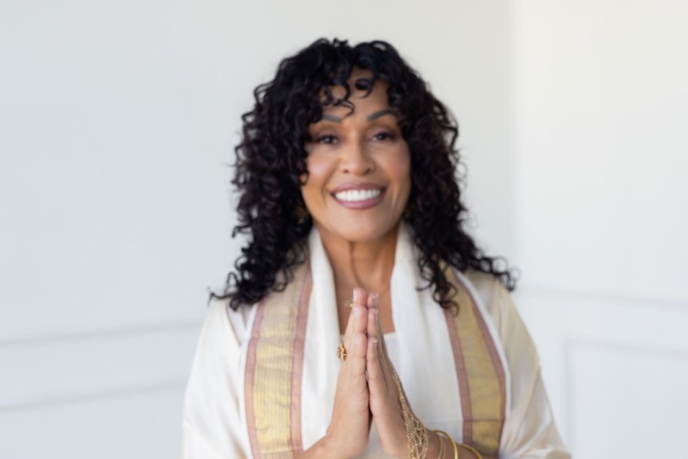 Maya Jaguar, an Interfaith Reverend and creator of Love Church, smiling with hands pressed together in a prayer pose, wearing traditional clothing, indoors with a white background.
