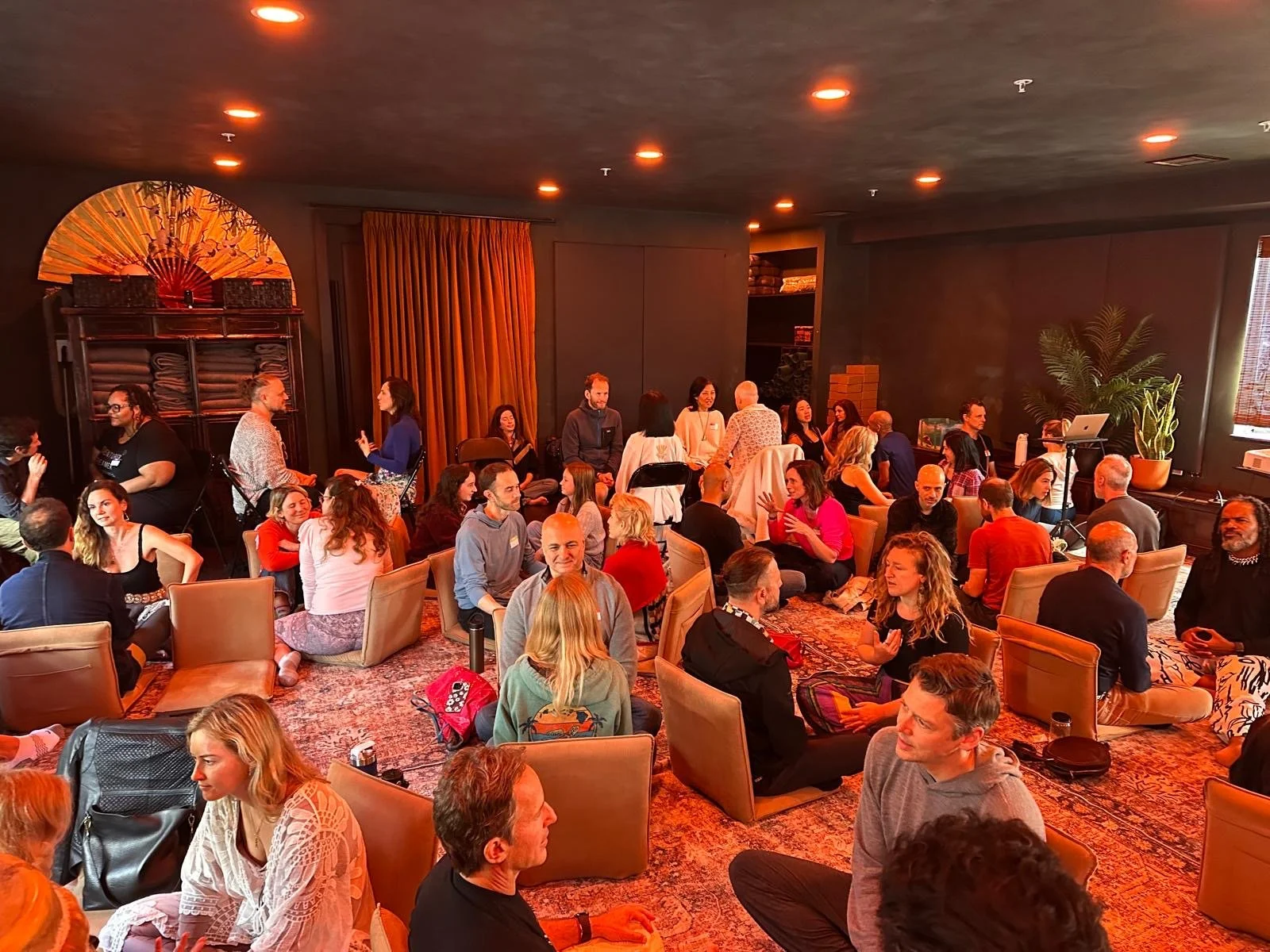 A large group of people gathered at Love Church in a cozy indoor setting, socializing and sitting on chairs and cushions on a patterned rug, with warm lighting, a plant, and a window with blinds in the background.