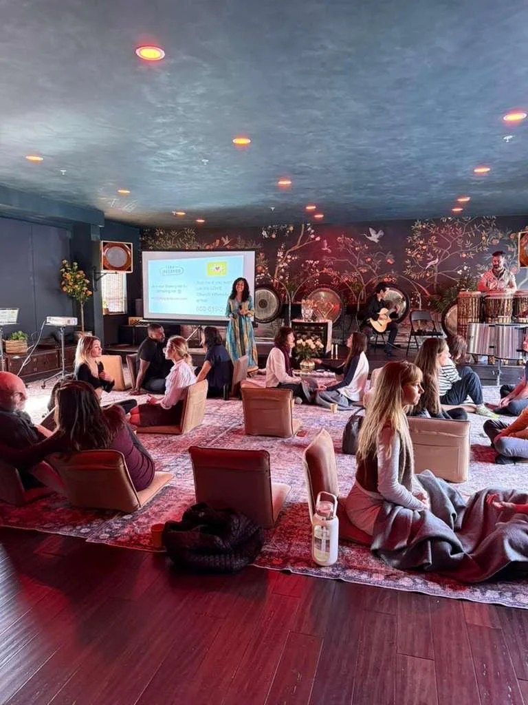 People sitting on cushions and chairs on a carpeted floor in a room with a musical performance. A woman is standing and speaking, with a large screen behind her displaying a presentation. There are musicians with instruments on a stage to the right, and the room has decorative floral wallpaper and hanging lights.