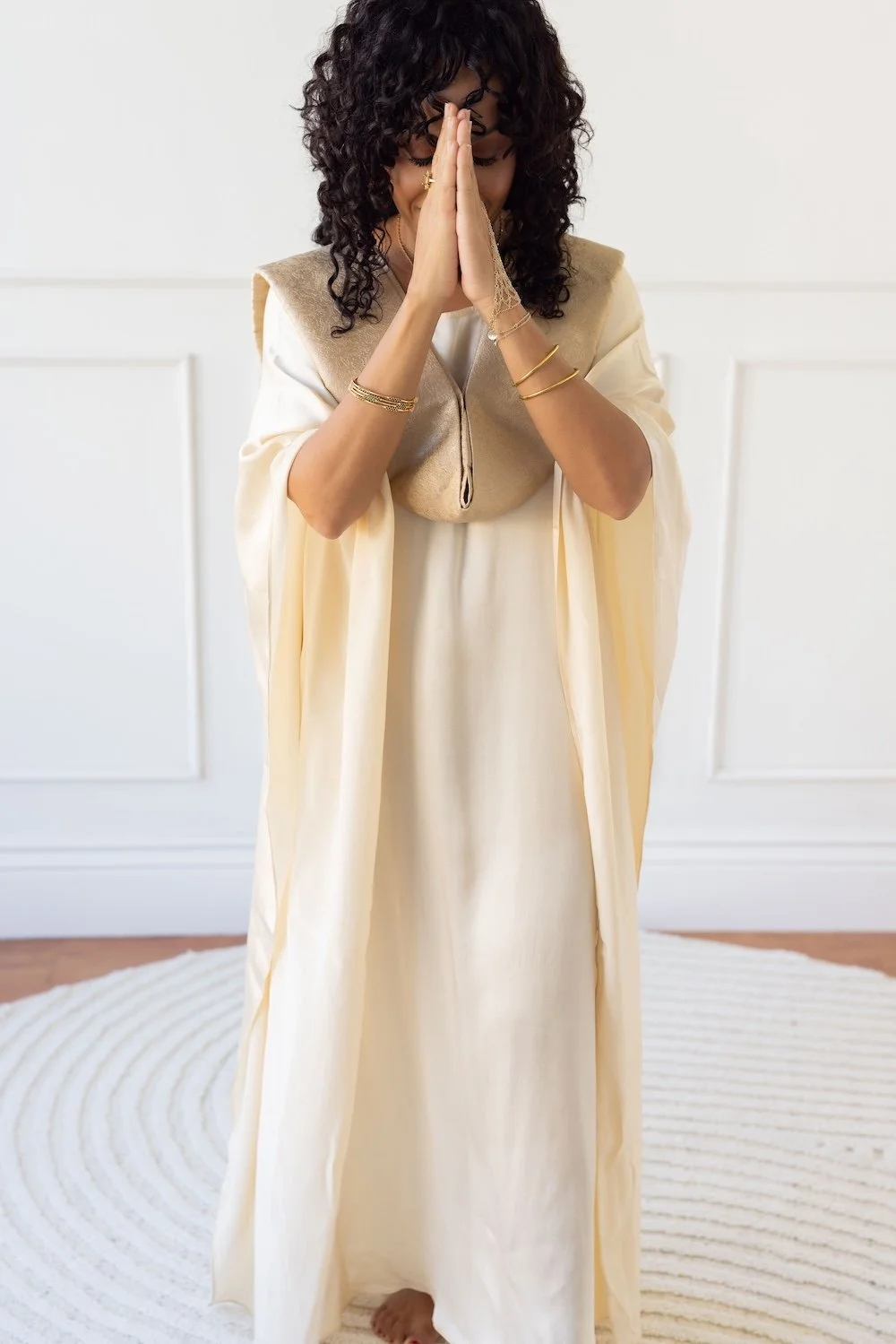 Maya Jaguar, an interfaith reverend, standing with her hands pressed together in prayer, wearing a flowy cream-colored dress and gold jewelry, in a bright room with white walls and a cream rug.