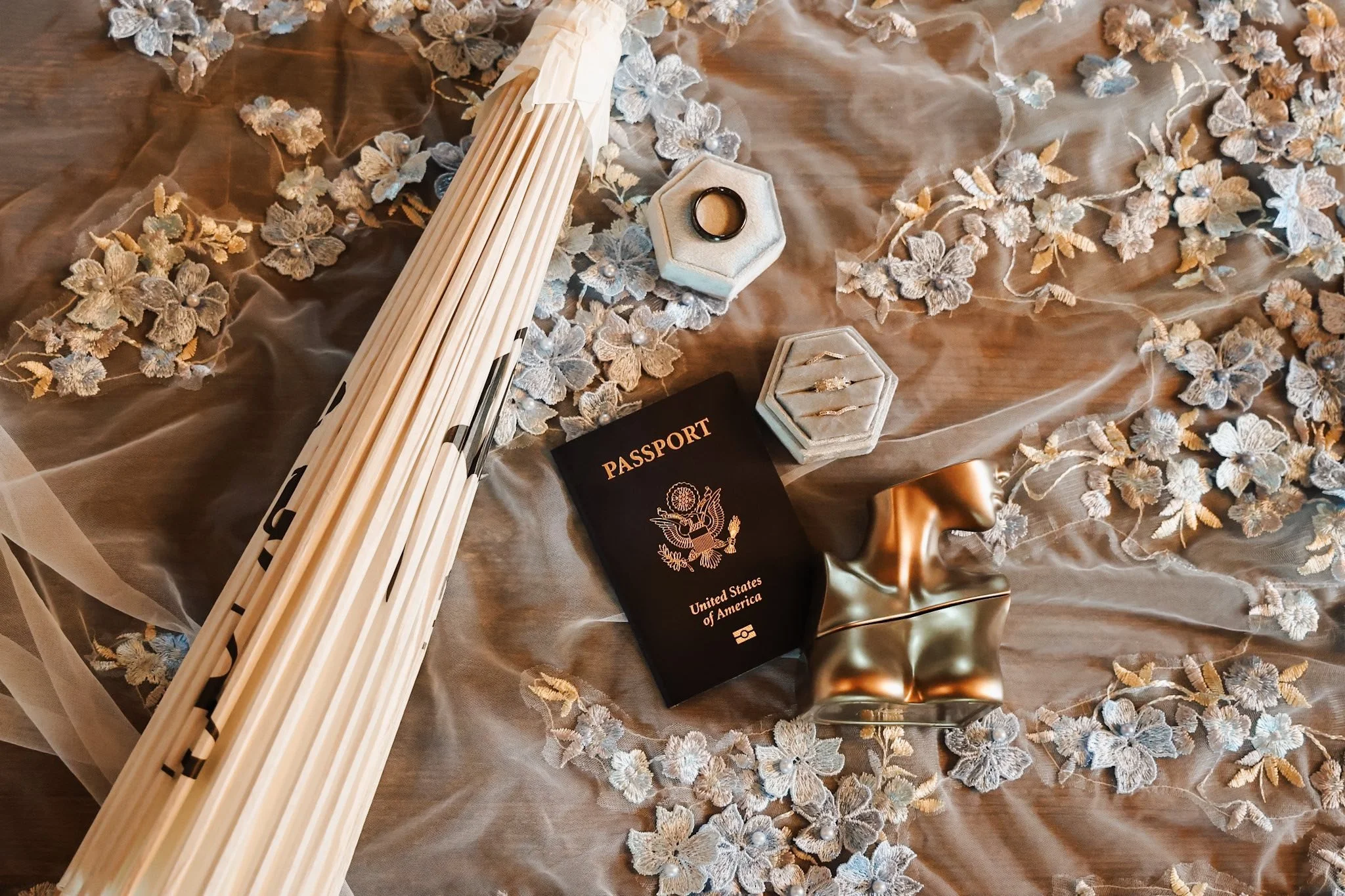 Wedding decorations including a bouquet, wedding rings, a passport, a gold dress, and decorative lace on a silky fabric background.