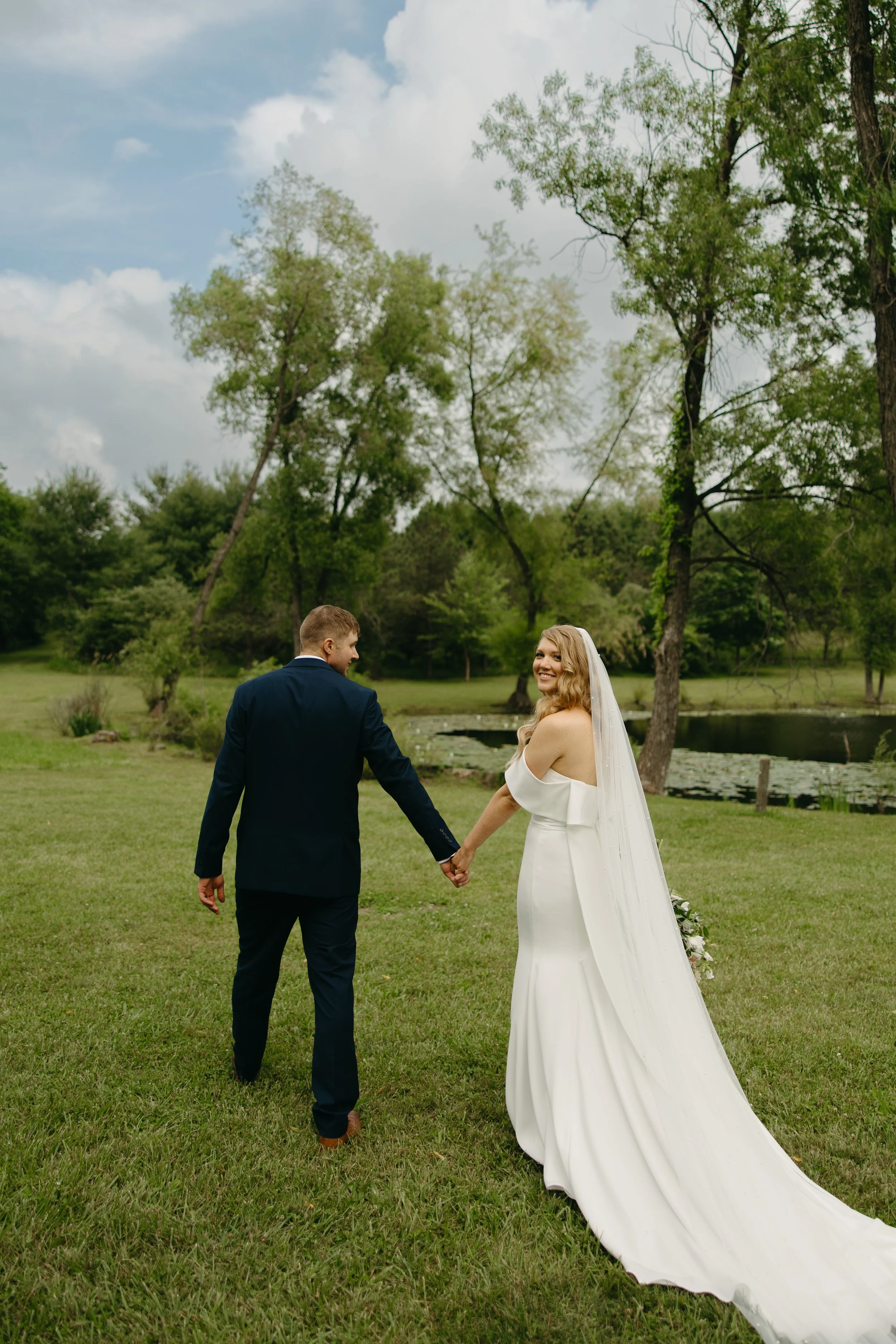 A bride and groom holding hands, walking outdoors on a grassy area near a pond, surrounded by trees during daytime.