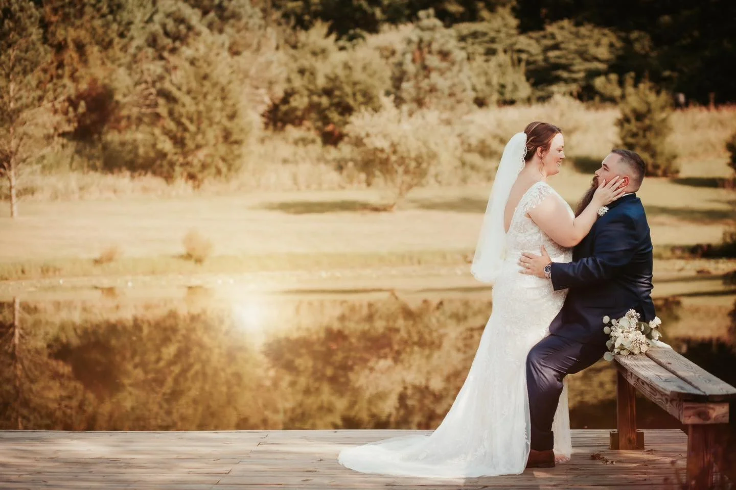A bride and groom embrace on a wooden dock by a lake during sunset, with trees and autumn foliage in the background.