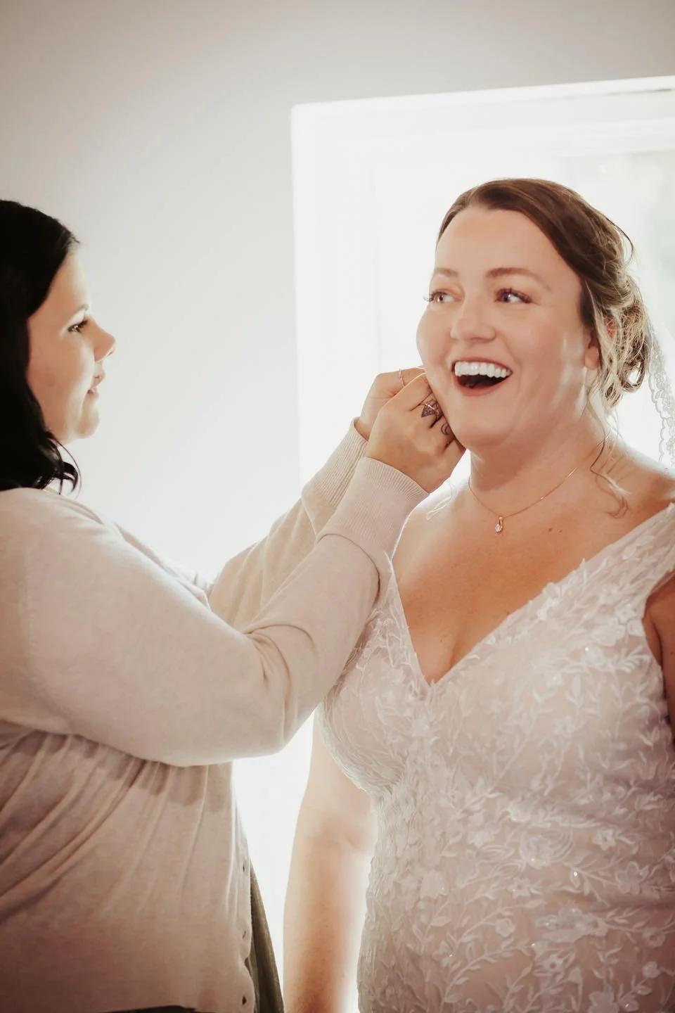 A woman in a bridal gown smiling as another woman helps her with earrings in a bright room.