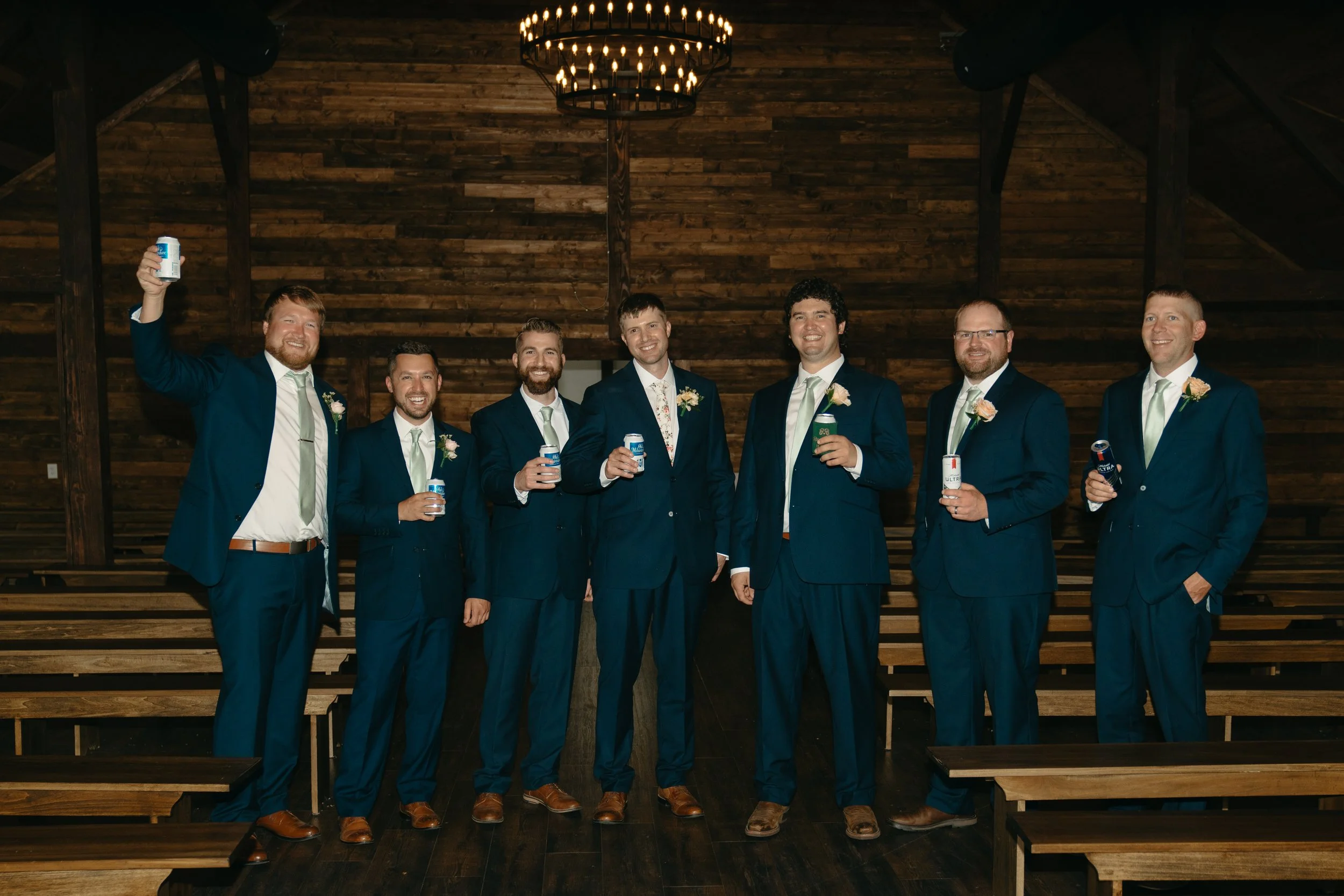 A group of eight men dressed in matching blue suits, white shirts, and light-colored ties, standing inside a wooden chapel holding cans of beverages, smiling at the camera. They are arranged in a semi-circle, with a wooden wall and chandelier behind 