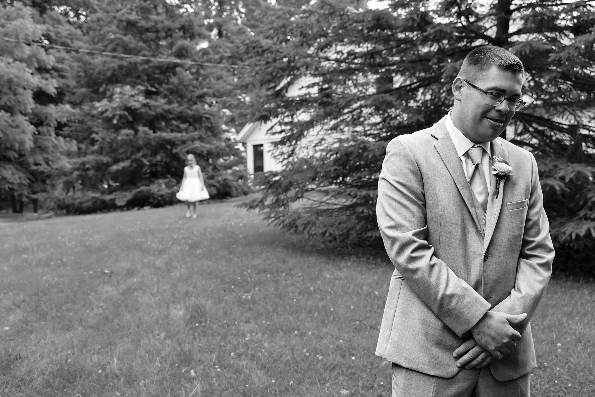 A man in a suit standing outdoors with a woman in a dress walking in the background among trees and grass.
