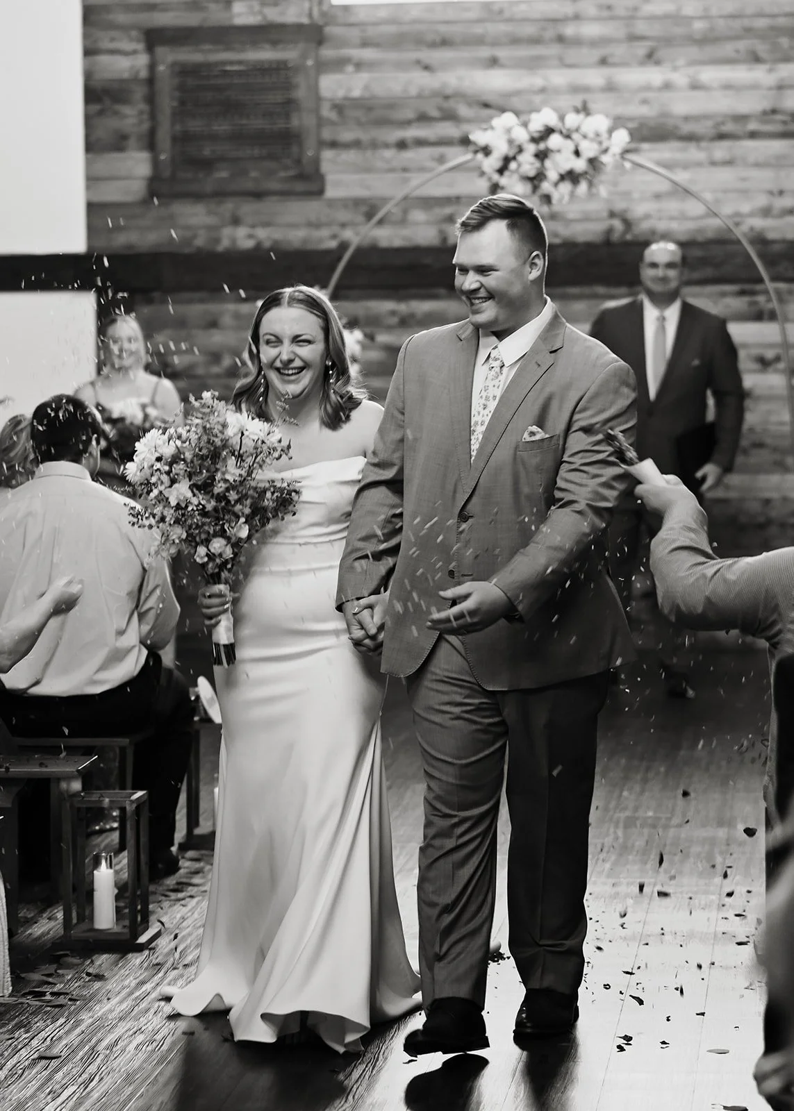 A black and white photo of a newlywed couple smiling, holding hands, walking down the aisle at their wedding reception, surrounded by guests throwing confetti.
