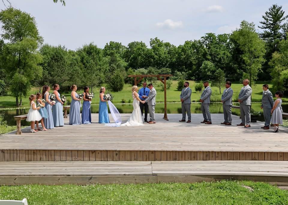 A wedding ceremony taking place outdoors on a wooden platform by a small lake, with trees in the background. The bride and groom are holding hands under a wooden arch. Bridesmaids and flower girls in blue dresses are standing on one side, holding bou
