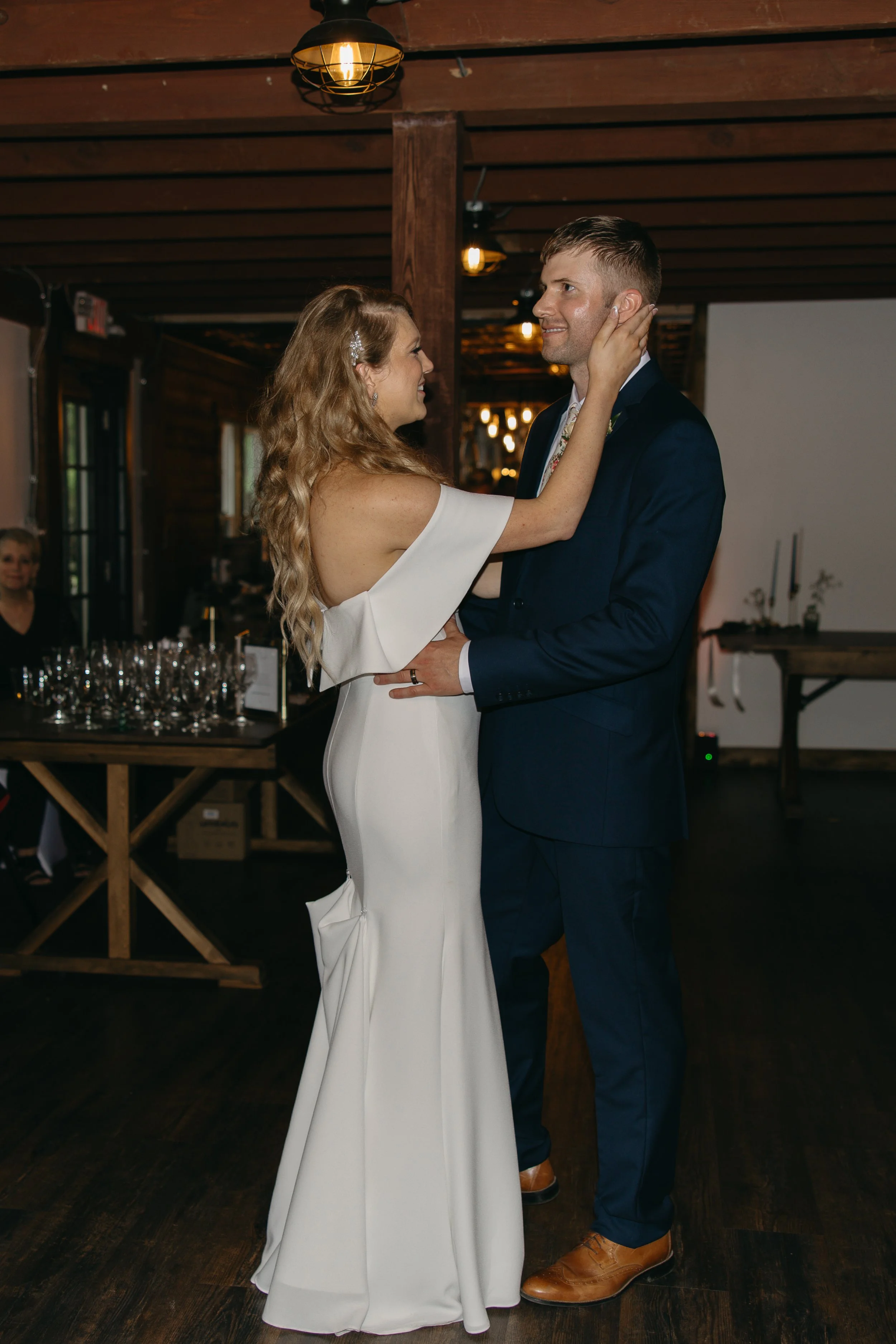 A bride and groom share a dance at their wedding reception in a rustic wooden venue. The bride has long, wavy blonde hair and is wearing a white, off-the-shoulder wedding dress. The groom is dressed in a navy suit and brown shoes. Soft, warm lighting