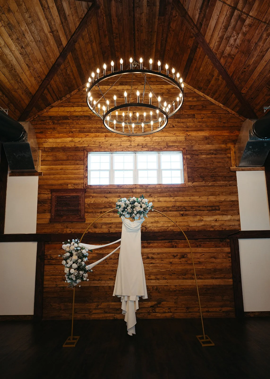 Indoor wedding altar with floral arrangements, set against a wooden wall with a window and a large circular chandelier hanging from the ceiling.