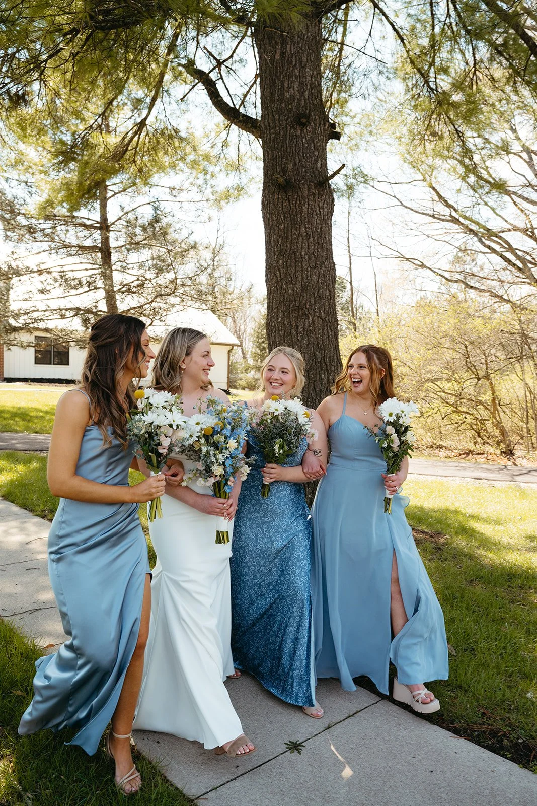 Four women in colorful dresses holding bouquets of flowers, walking and laughing outdoors under a large tree.