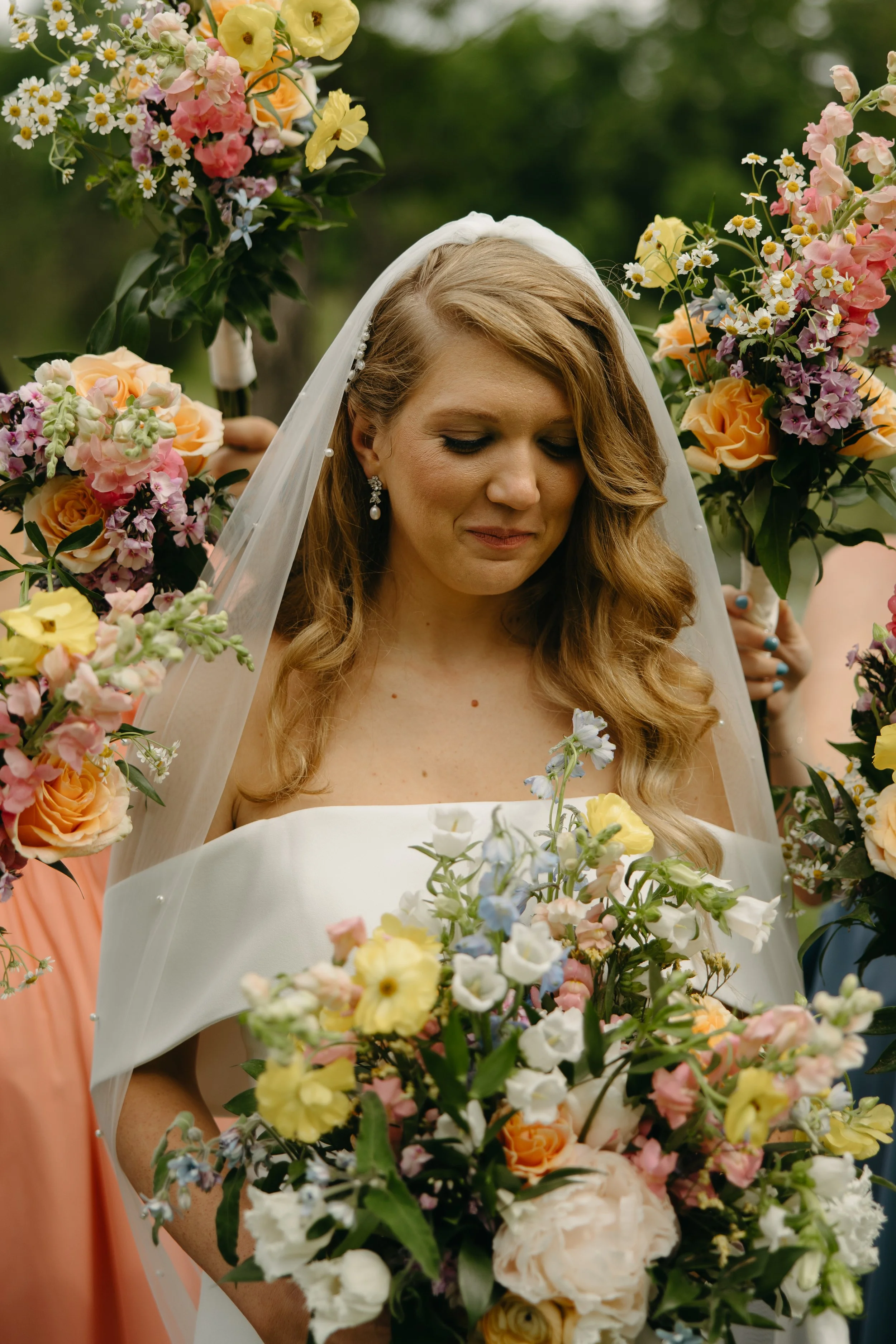 A bride with long, wavy, light brown hair stands outdoors, holding a bouquet of flowers. She wears a white strapless wedding dress, a sheer veil over her head, and elegant drop earrings. Surrounding her are bridesmaids holding large bouquets of color