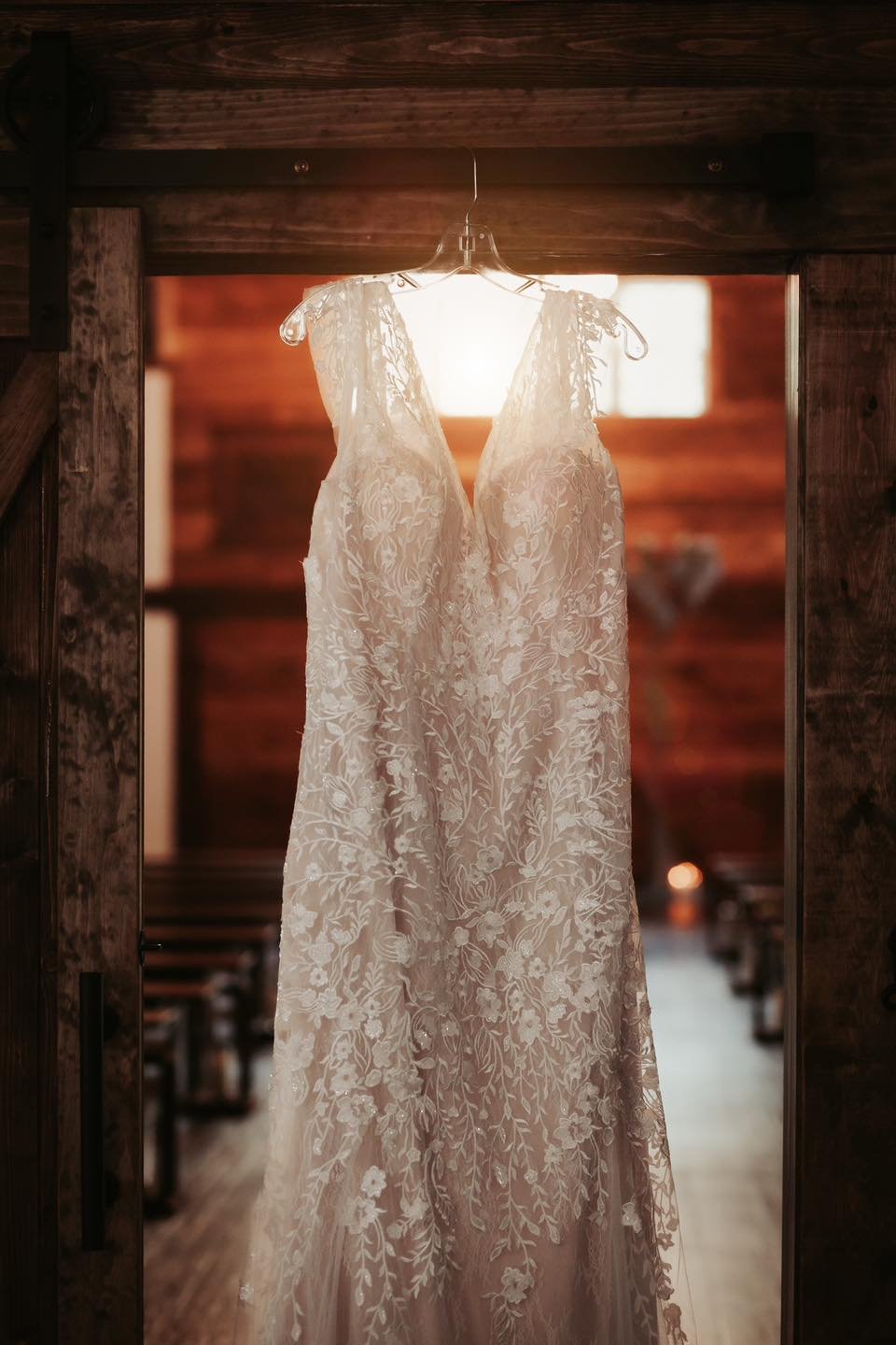 Elegant wedding dress with floral lace details hanging on a hanger in a rustic wooden interior.