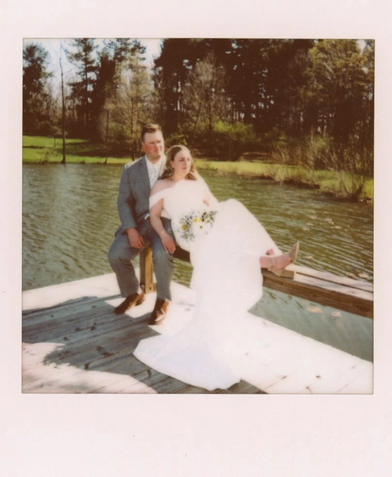 A bride and groom sitting on a dock by a lake, with trees in the background, during daytime.