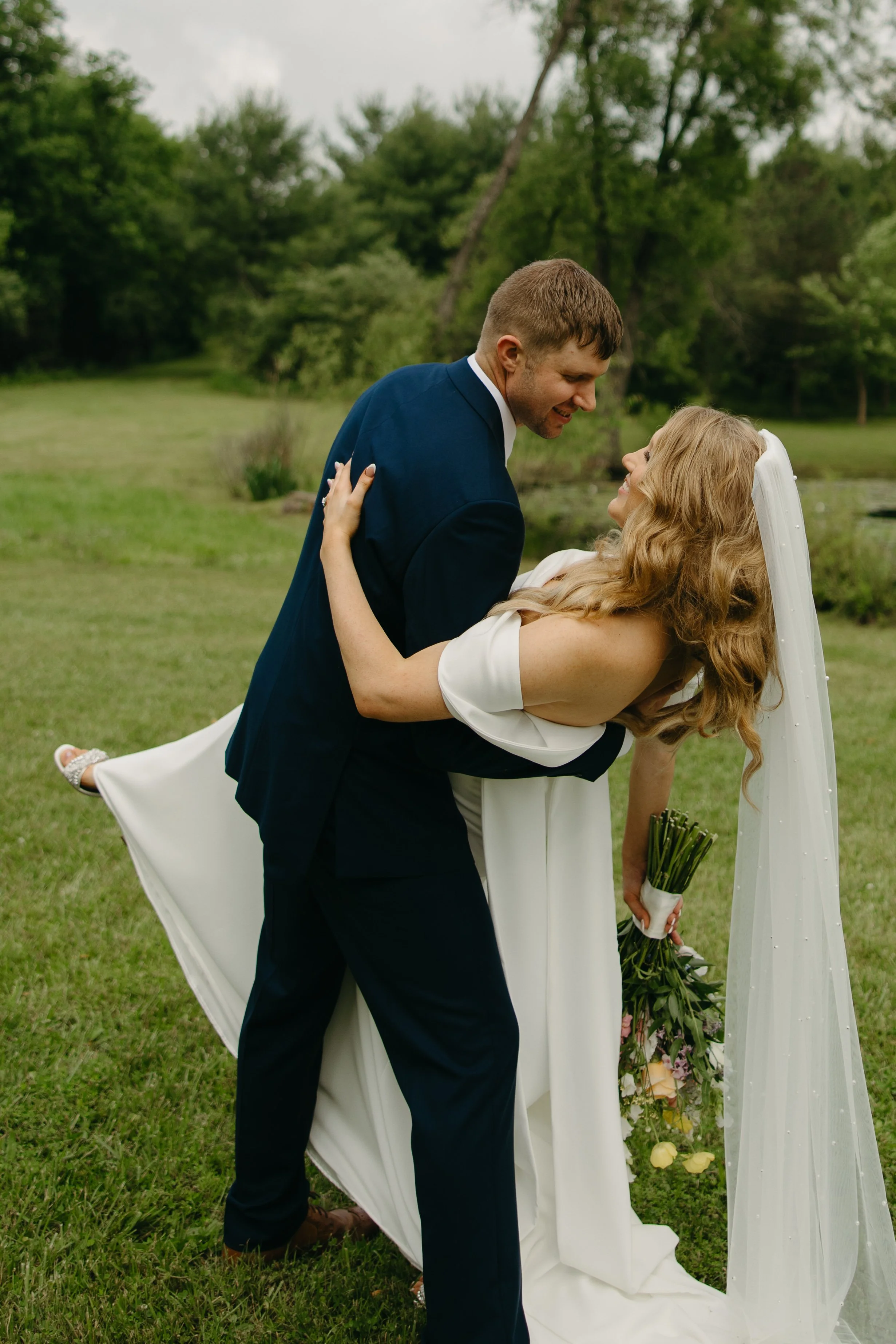 A newlywed couple outdoors on their wedding day, with the groom dipping the bride, who is holding a bouquet of flowers, in front of a green landscape with trees.