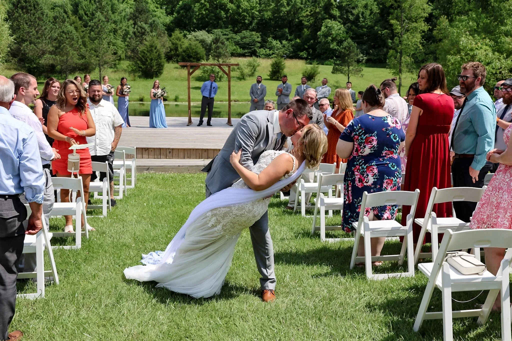 A newlywed couple kissing during their outdoor wedding ceremony. The groom dips the bride as she leans back, surrounded by seated and standing guests under a bright, sunny day with a grassy field and trees in the background.