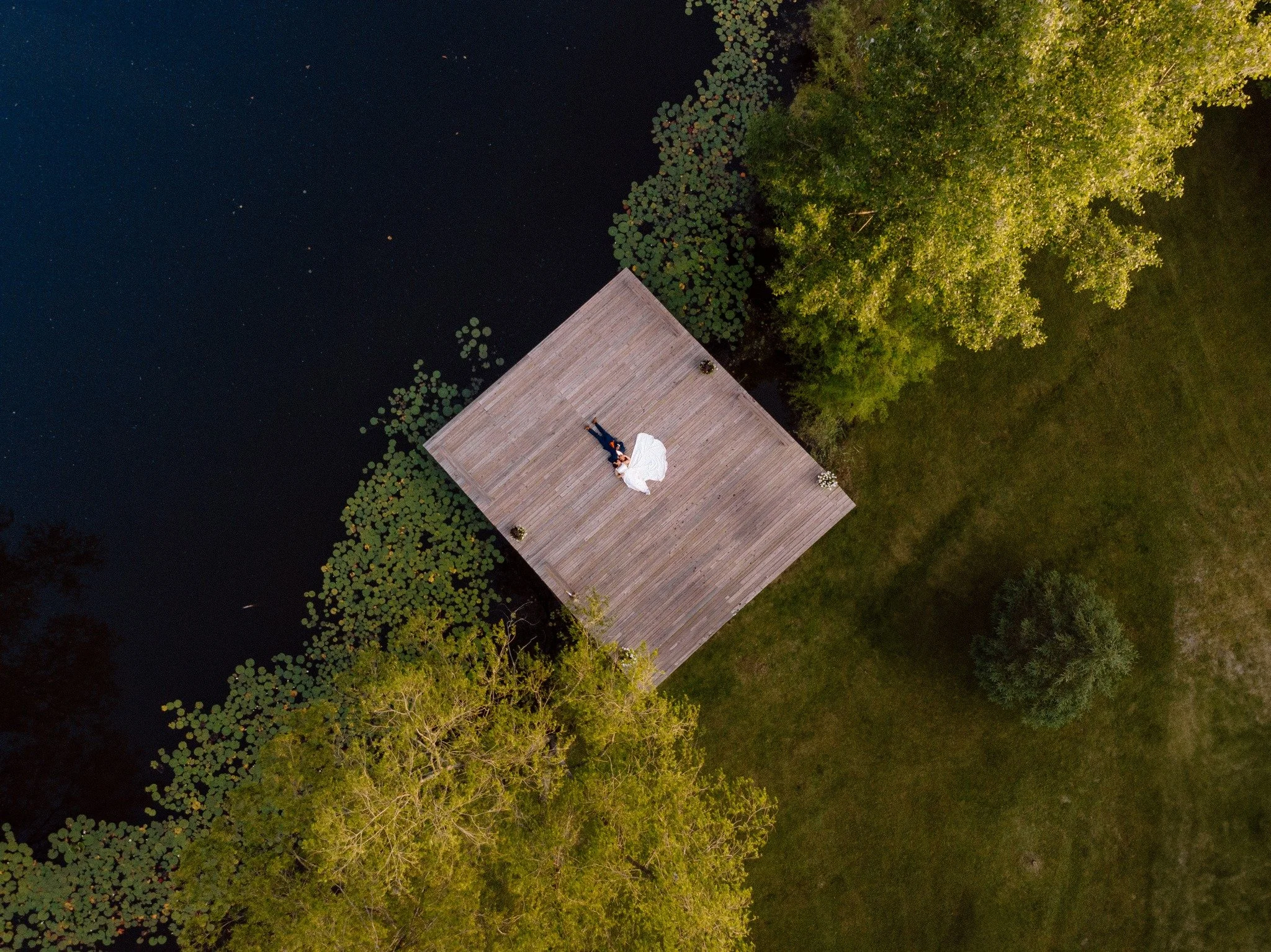 A couple lying on a wooden dock by a lake, with one person in a white dress and the other in a dark suit, surrounded by green trees at dusk.