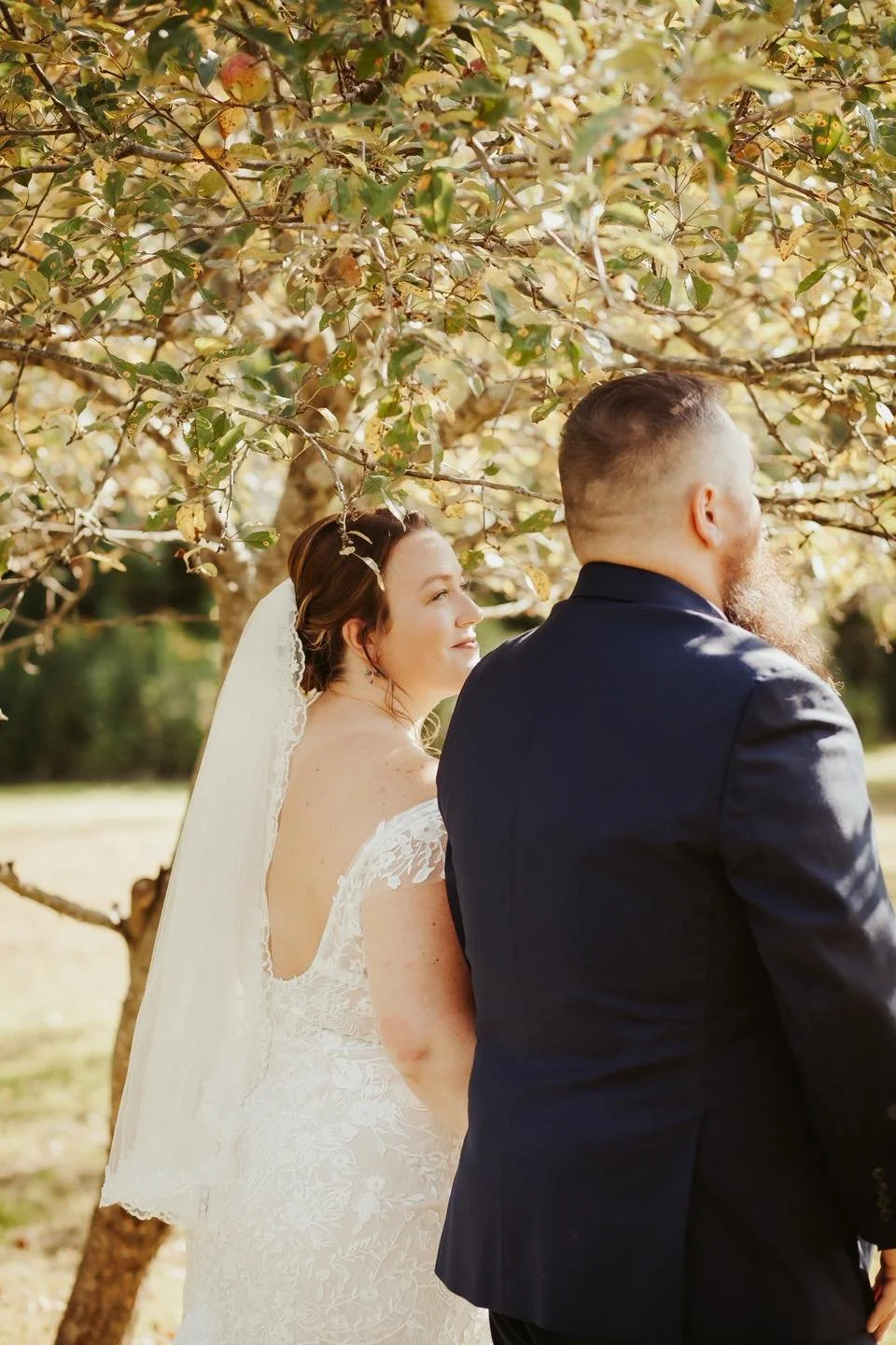 Bride and groom standing outdoors under a tree on their wedding day, facing away from the camera, with sunlight filtering through the leaves.