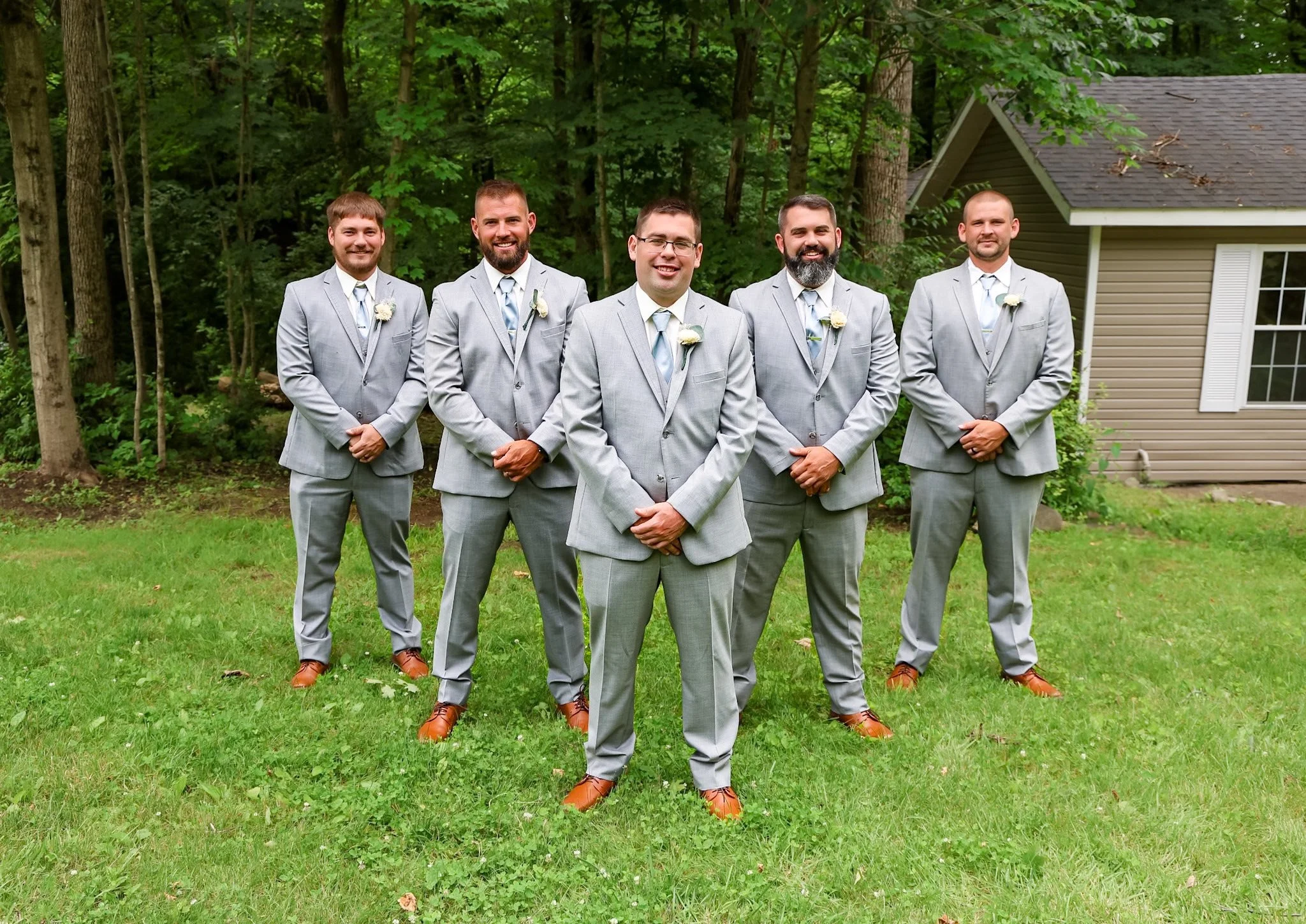 Six men in light gray suits with wedding boutonnieres pose outdoors on a grassy area with trees and a house in the background. They are smiling and standing in a semi-circle.