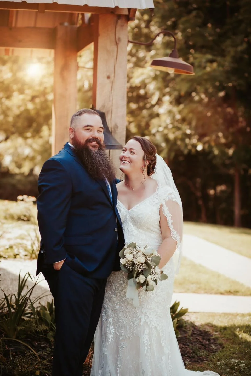Bride and groom standing outdoors on their wedding day, with the bride holding a bouquet of white flowers and greenery, under a wooden structure with outdoor lighting, in a natural setting with trees and sunlight.