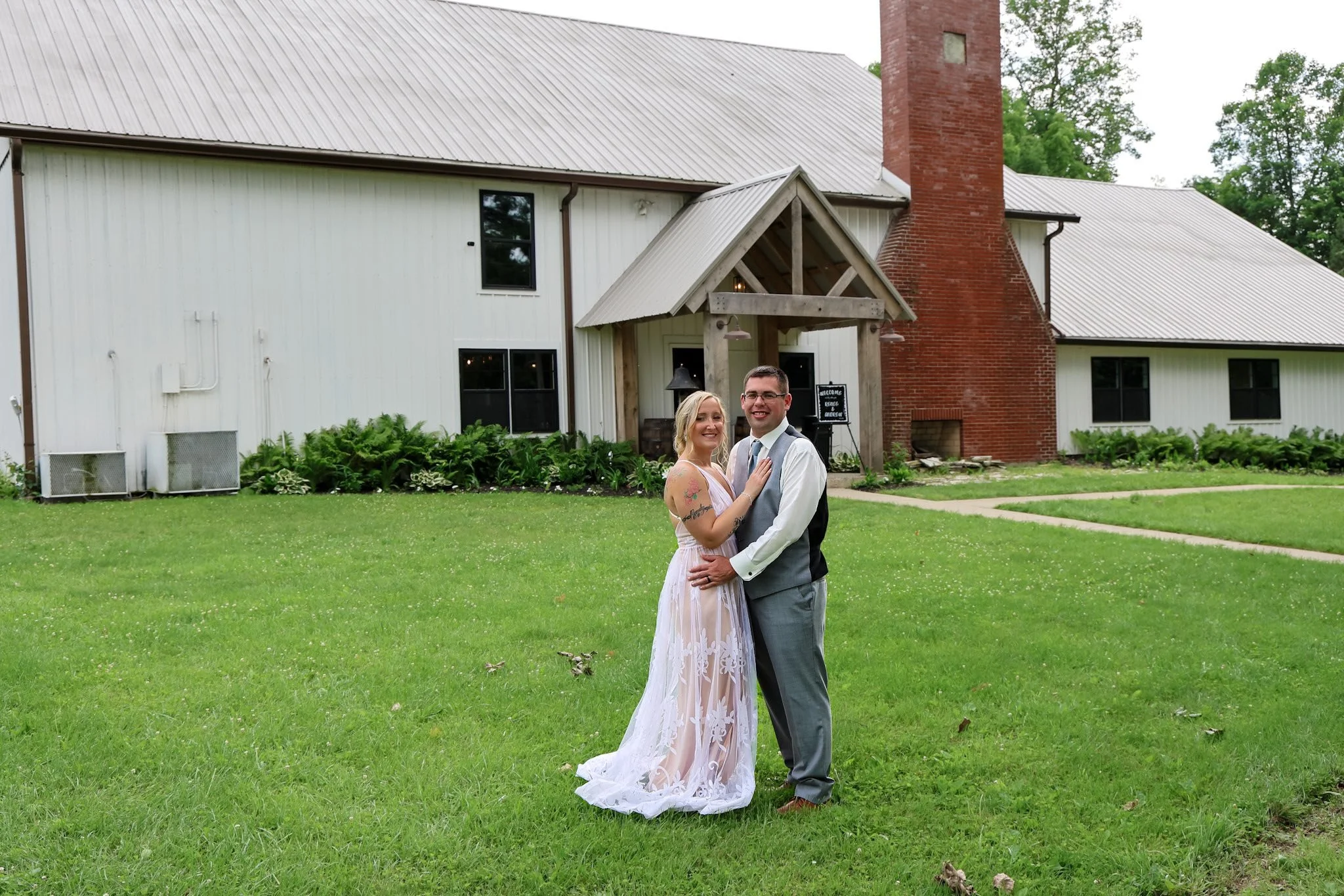 A couple in wedding attire standing on a green lawn in front of a white building with black windows and a red brick chimney, smiling and posing for a photo.
