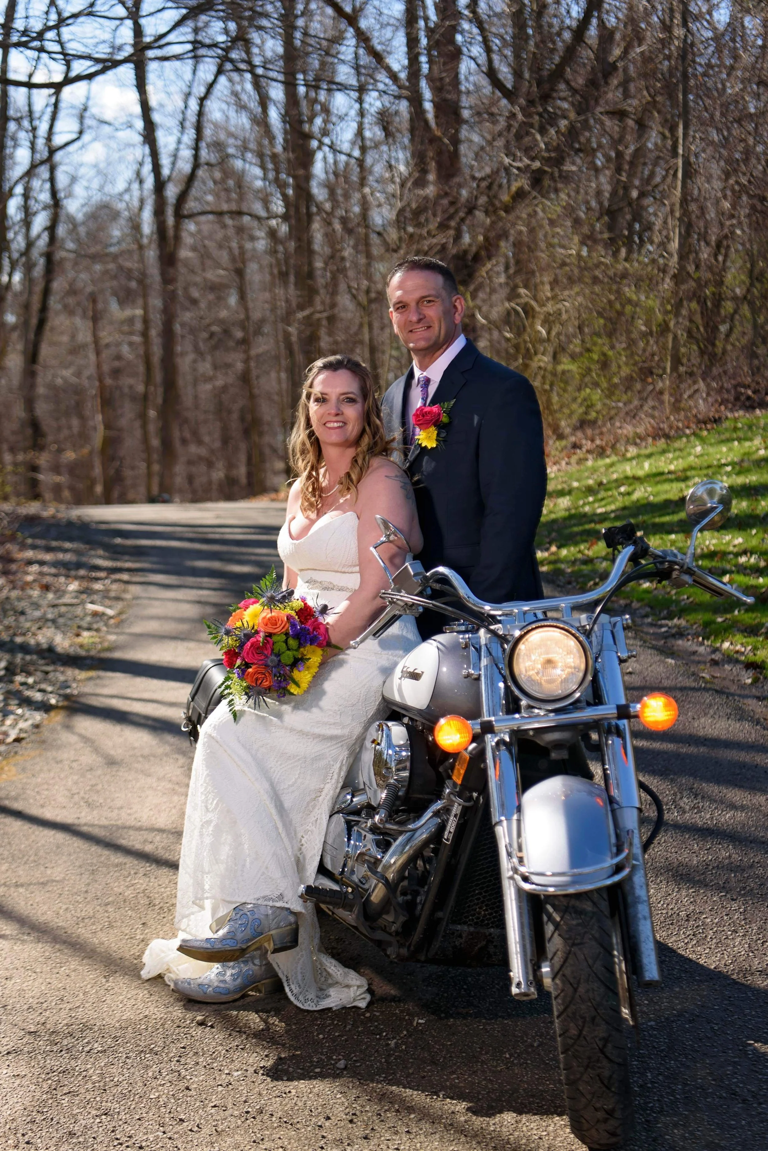 A bride and groom sitting on a motorcycle on a rural road during daytime, with leafless trees and grass in the background.