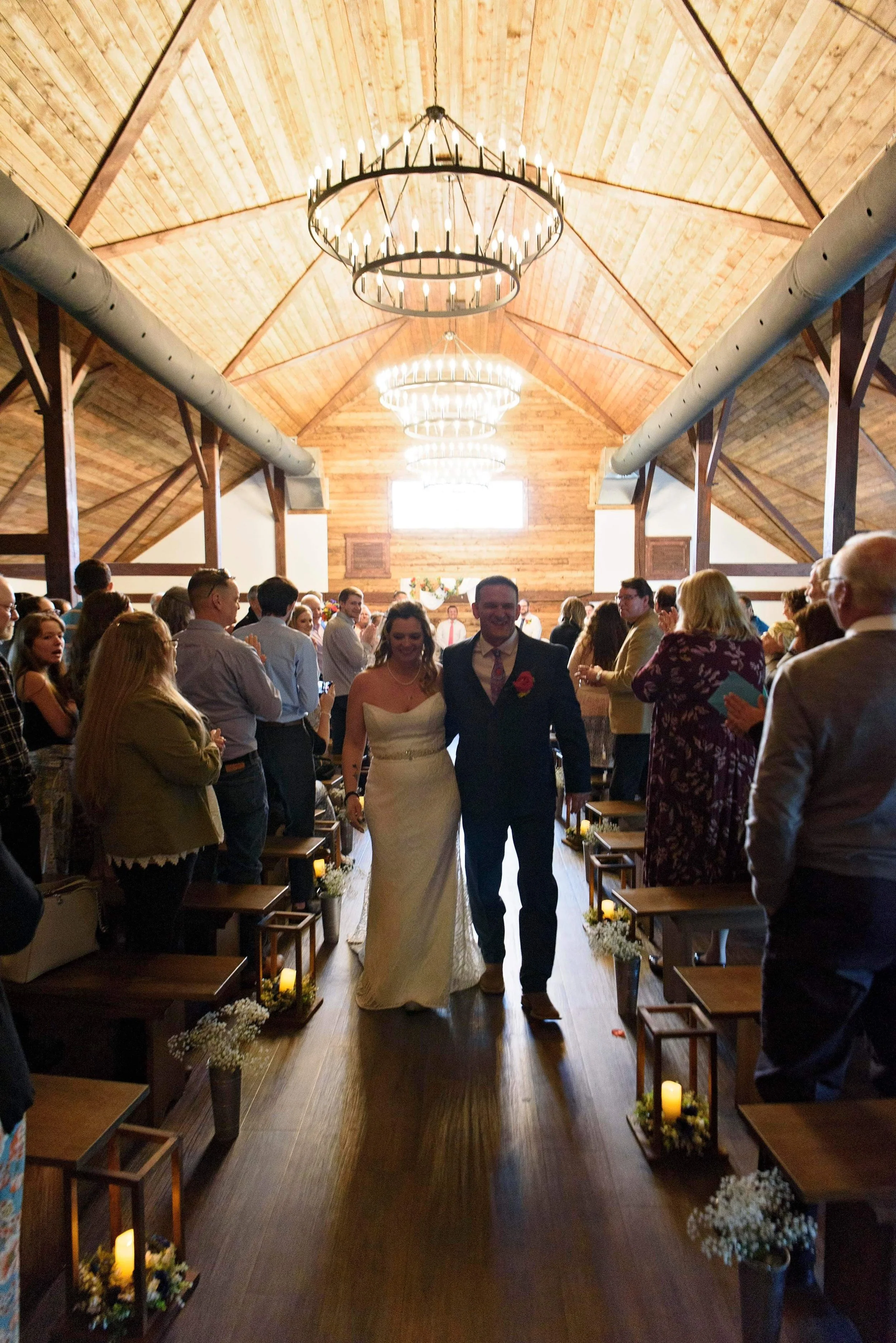 A bride and groom walking down the aisle at a wedding ceremony in a wooden, barn-like venue with guests on either side clapping and taking photos.