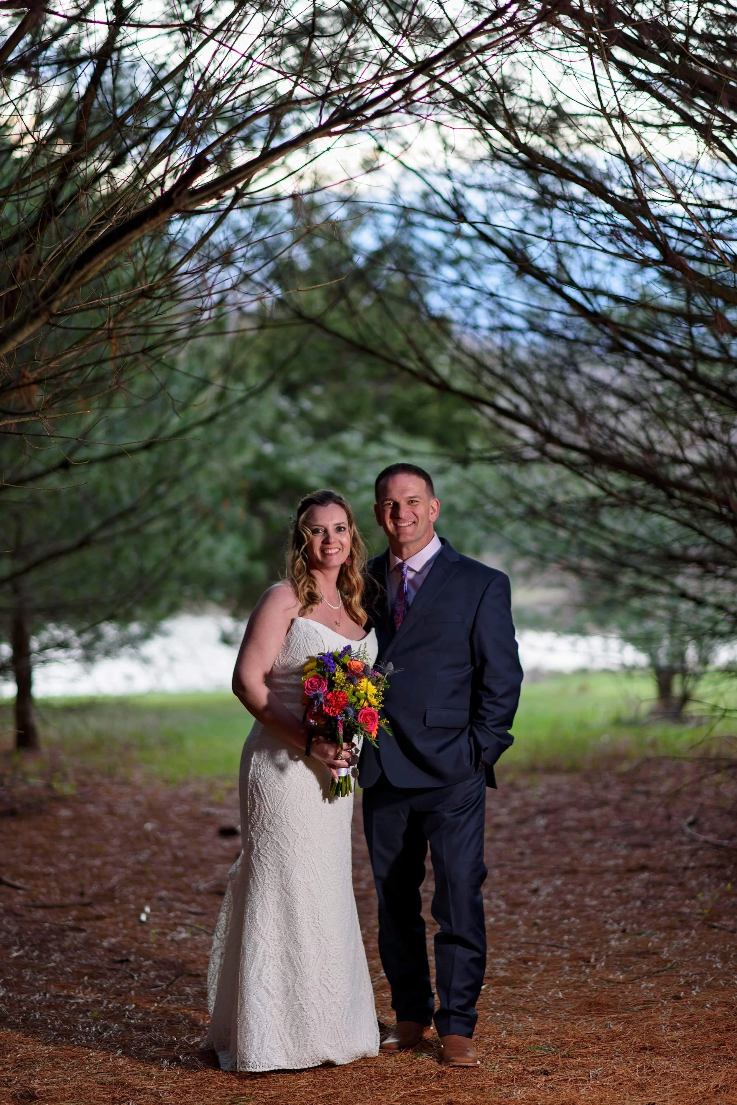 A bride wearing a white wedding gown and a groom in a dark suit pose outdoors under a canopy of trees, with the bride holding a bouquet of colorful flowers.