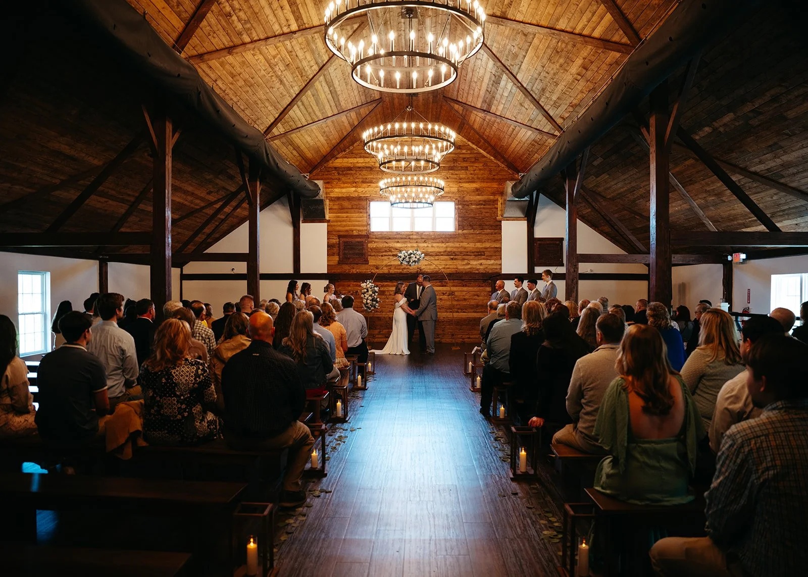 A wedding ceremony taking place inside a barn with wooden walls and ceiling, featuring chandeliers and a large window behind the altar. The bride and groom are standing at the altar with officiants and bridal party members present, surrounded by seat