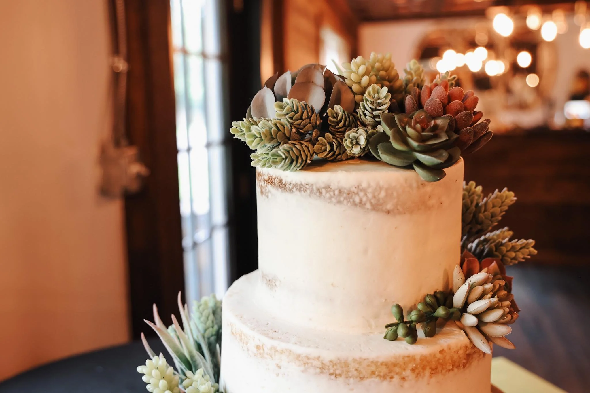 A two-tiered wedding cake decorated with succulents on top and cascading down the side, set in a warmly lit room with wooden walls and large windows.