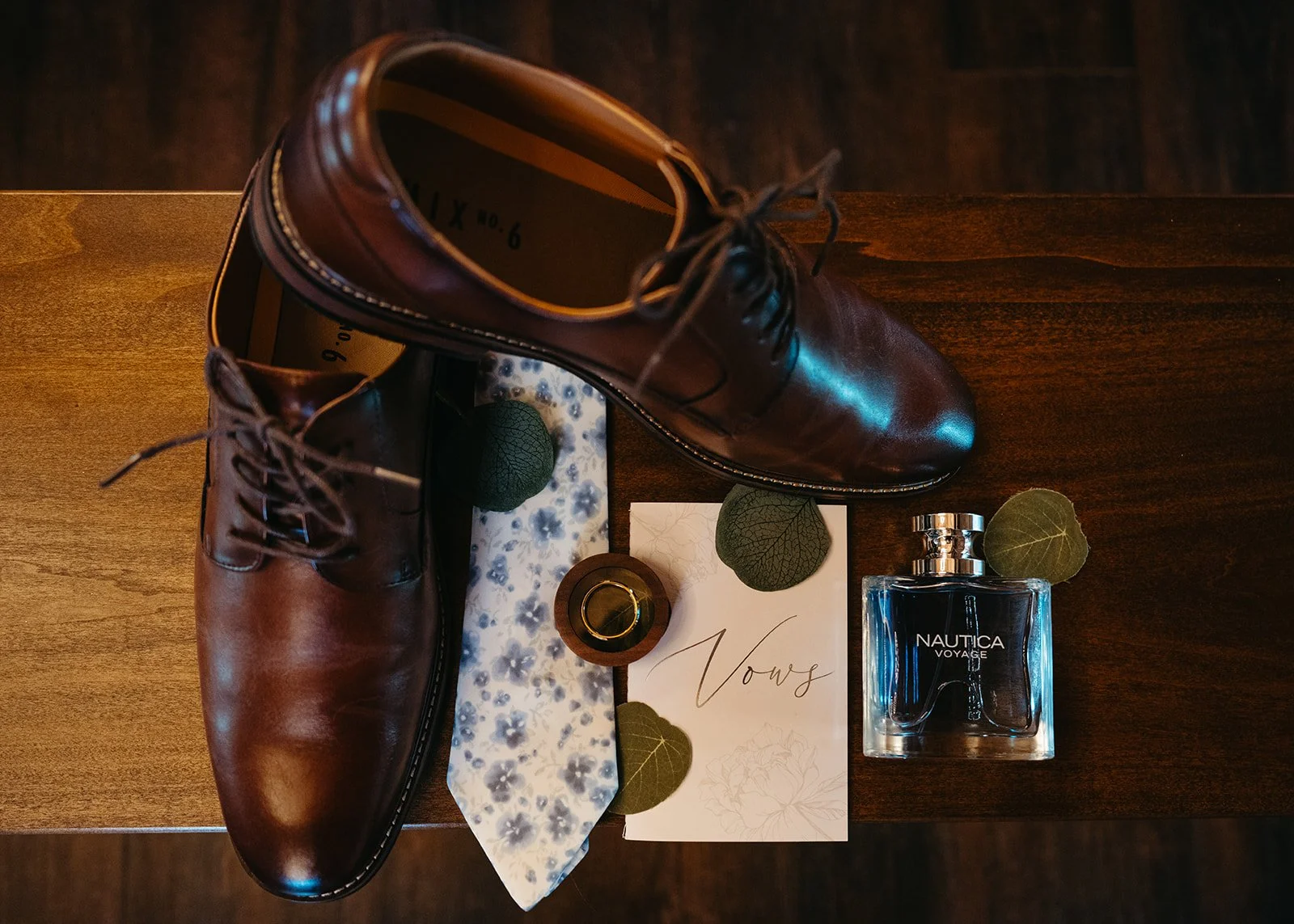 Top view of a pair of brown leather dress shoes, a perfume bottle labeled 'Nautica Voyage,' a wedding ring, a floral pocket square, a wedding invitation with the word 'Vow,' and some green leaves on a wooden surface.