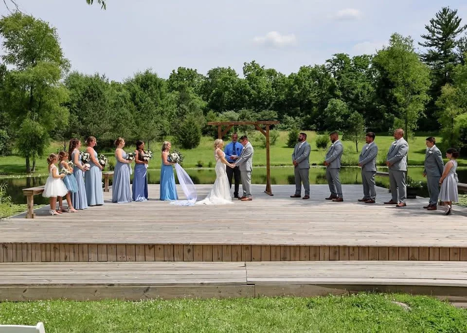 Outdoor wedding ceremony on a wooden platform by a pond with a green landscape and trees in the background, with the bride and groom at the center, surrounded by bridesmaids and groomsmen.