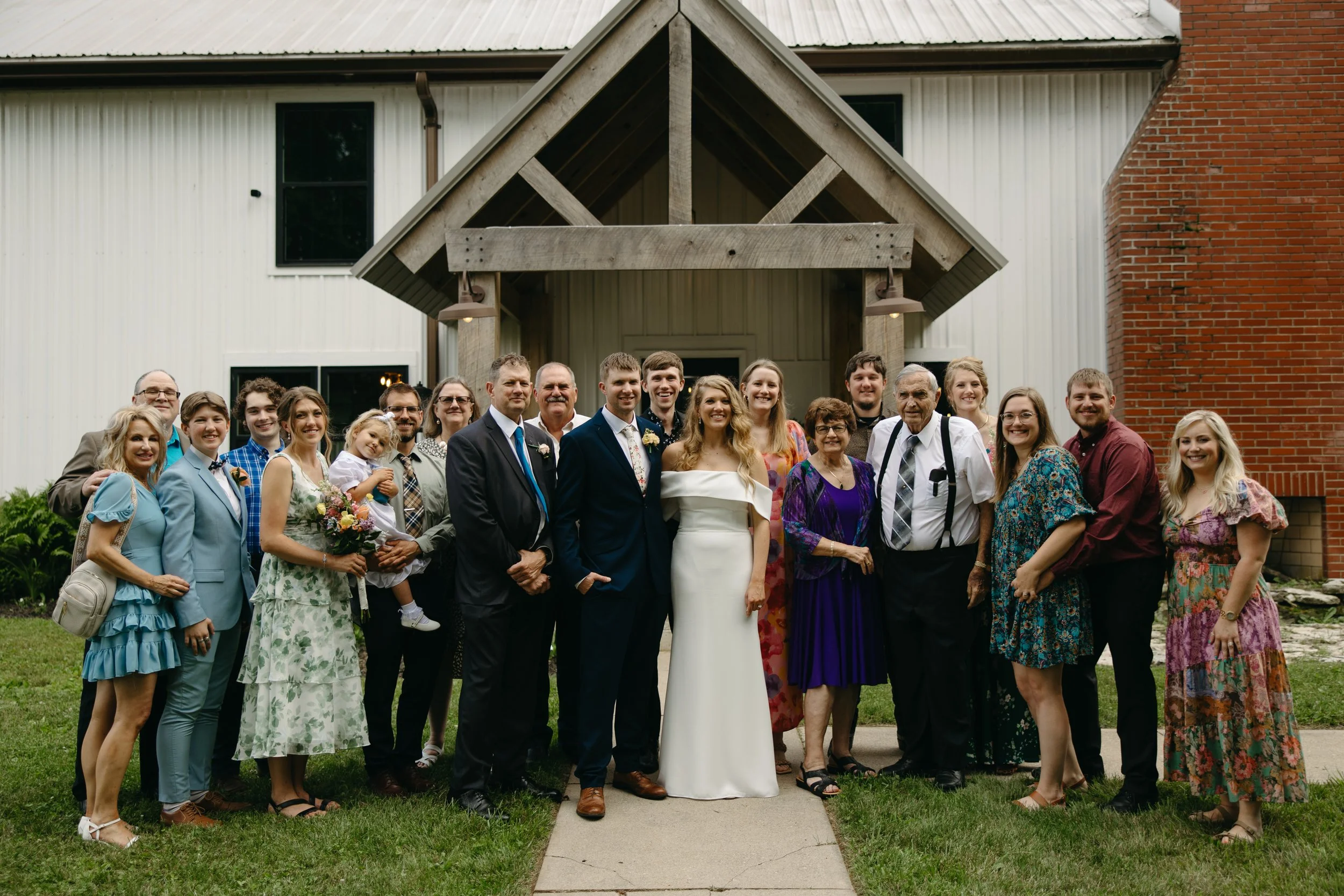 A group of people gathered outside a building for a wedding photo, including the bride and groom, family members, and friends, standing on a sidewalk in front of the building's entrance.