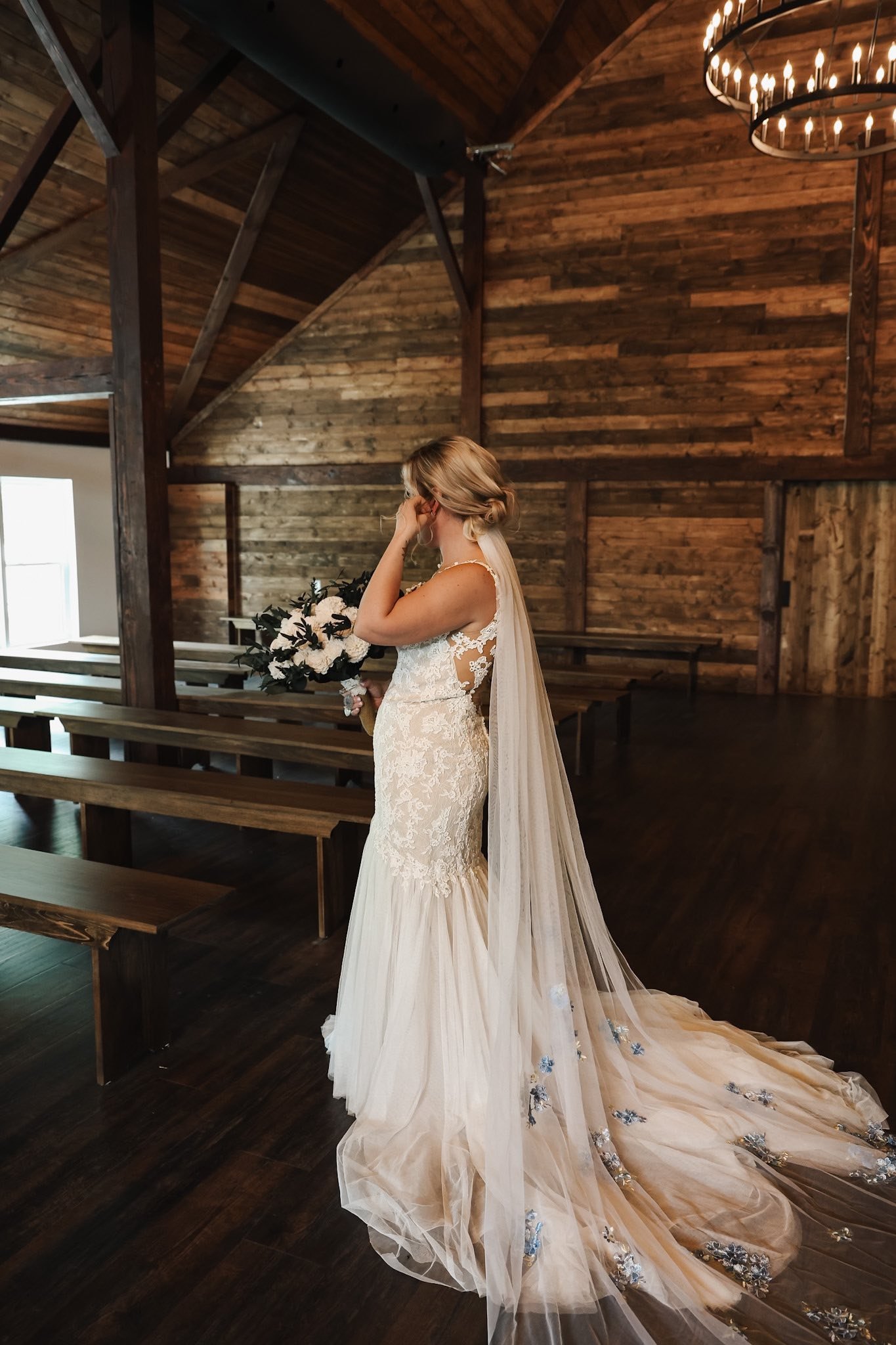 A bride in a lace wedding dress with a long tulle train and veil, holding a bouquet of flowers, standing indoors in front of wooden walls and benches, covering her face with her hand during a moment of emotion.