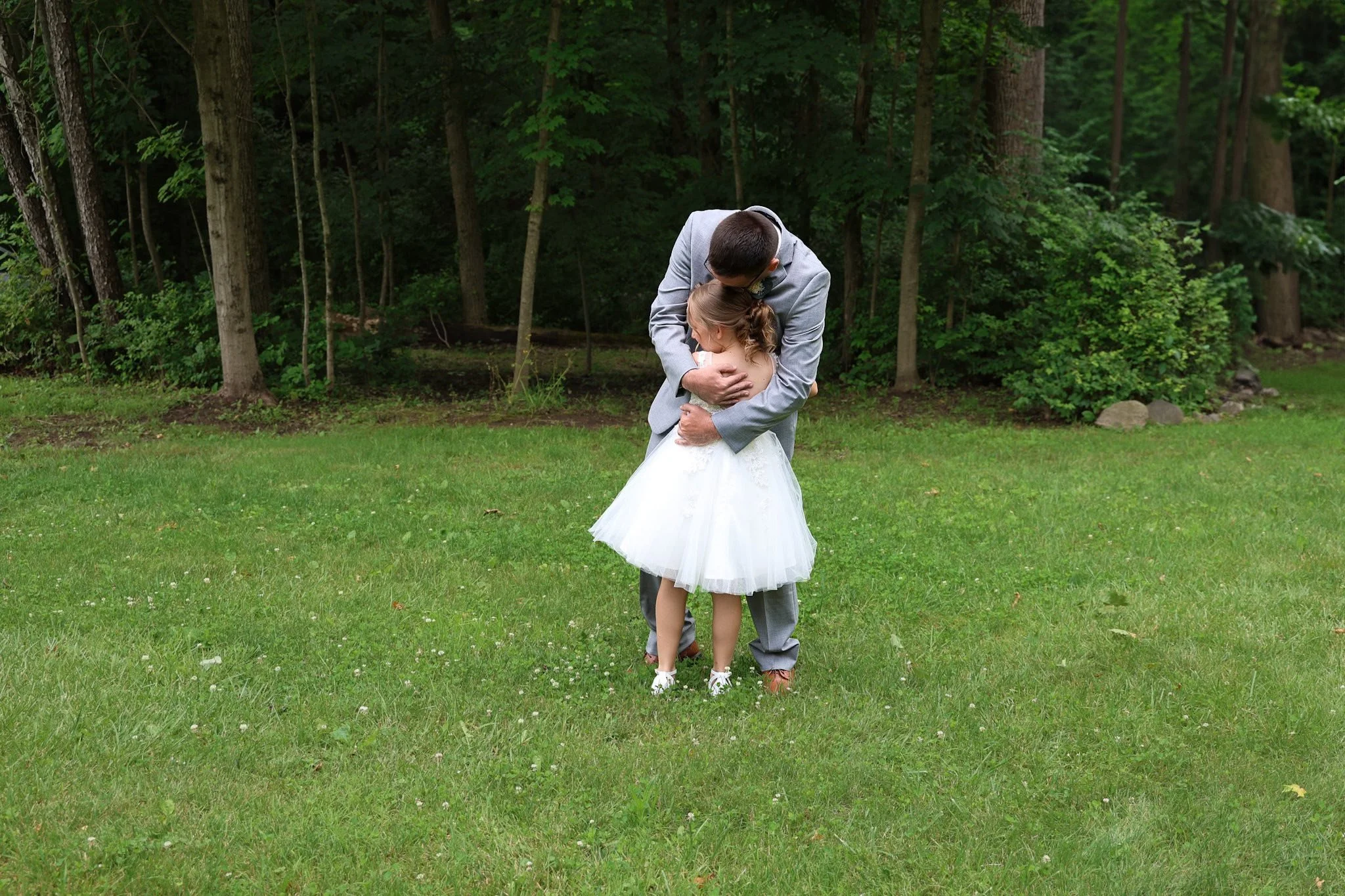 A man in a gray suit hugging a young girl in a white dress in a grassy field with trees in the background.
