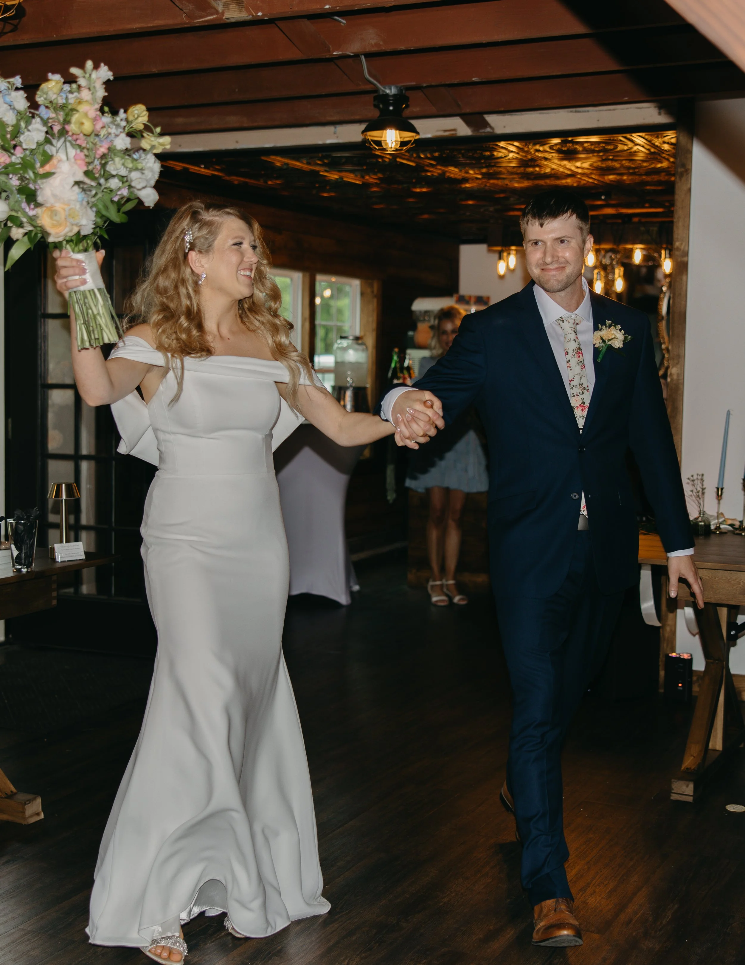 A bride and groom holding hands and walking inside a rustic wedding venue with wooden walls and ceiling, smiling and enjoying their special moment.