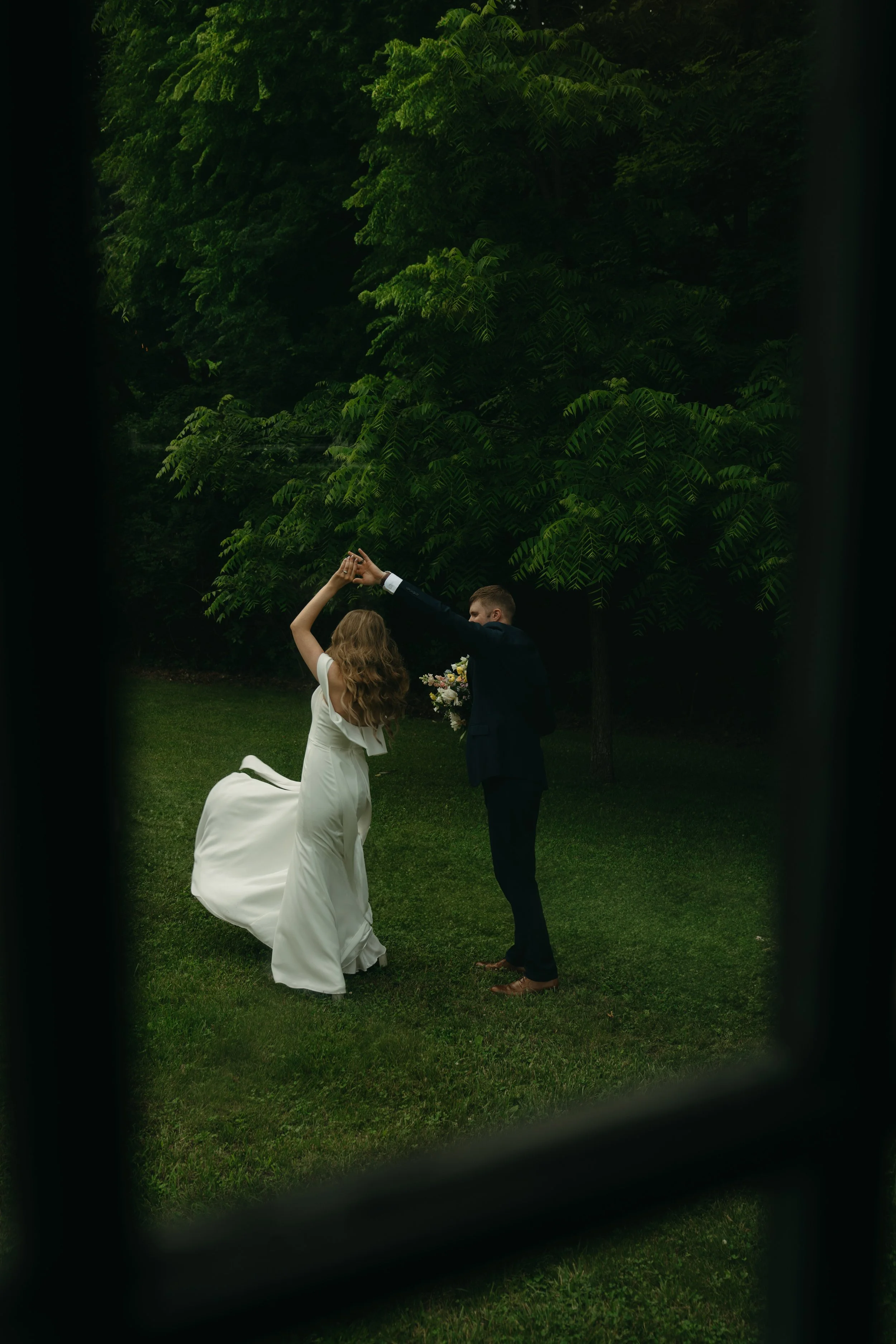 A bride and groom dancing outdoors on a grassy area surrounded by trees, viewed through window panes.