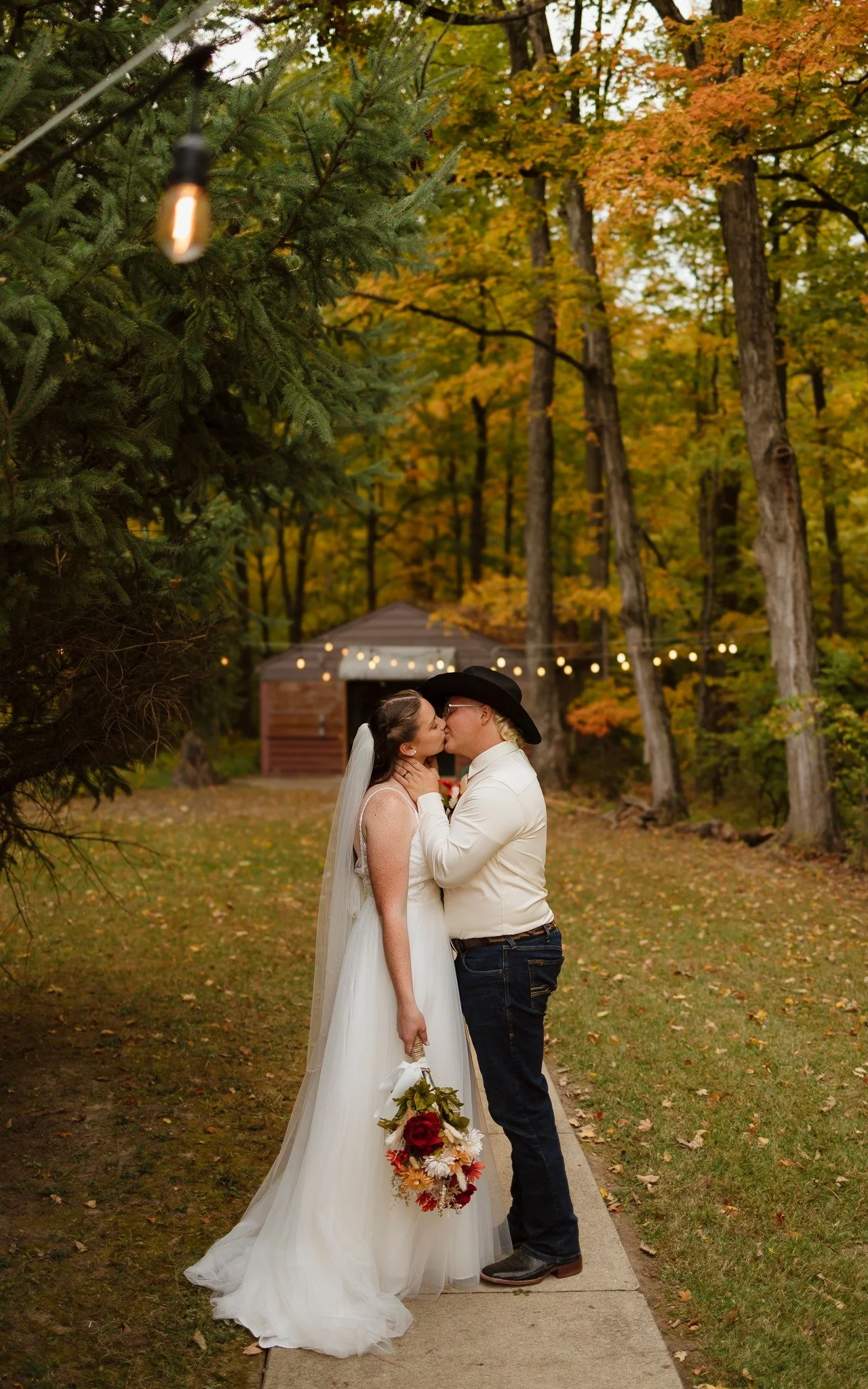 A bride and groom sharing a kiss outdoors during their wedding, with fall foliage and string lights in the background.
