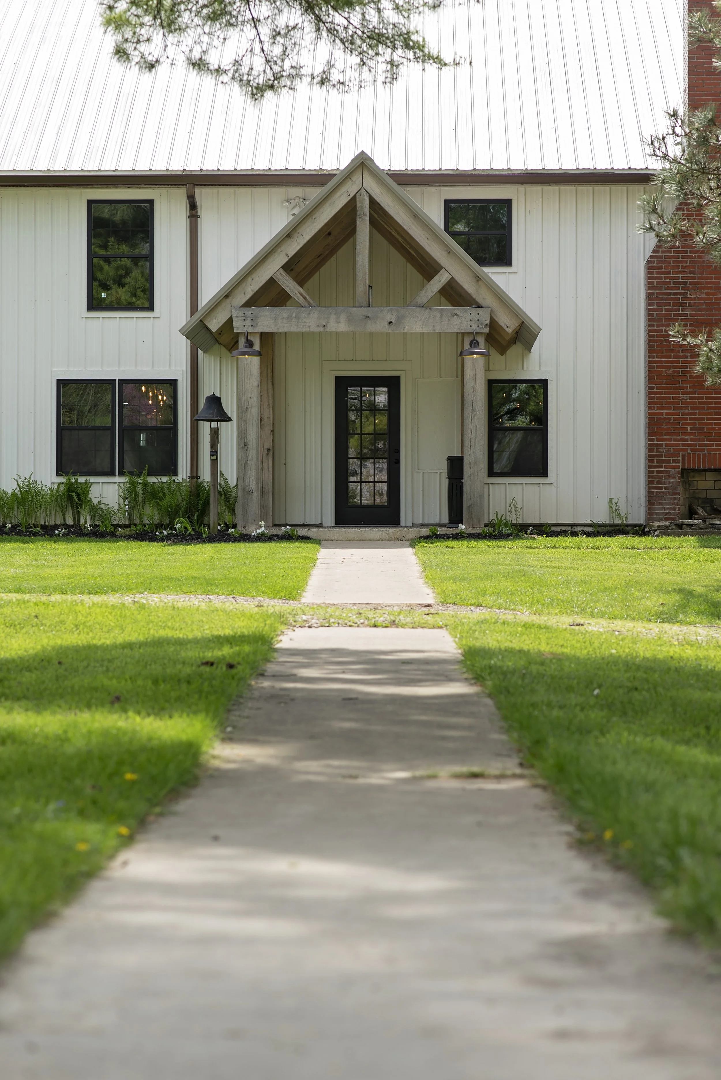 Front view of a modern house with a concrete walkway leading to the black front door, green lawn on either side, and a small wooden porch with outdoor lighting.