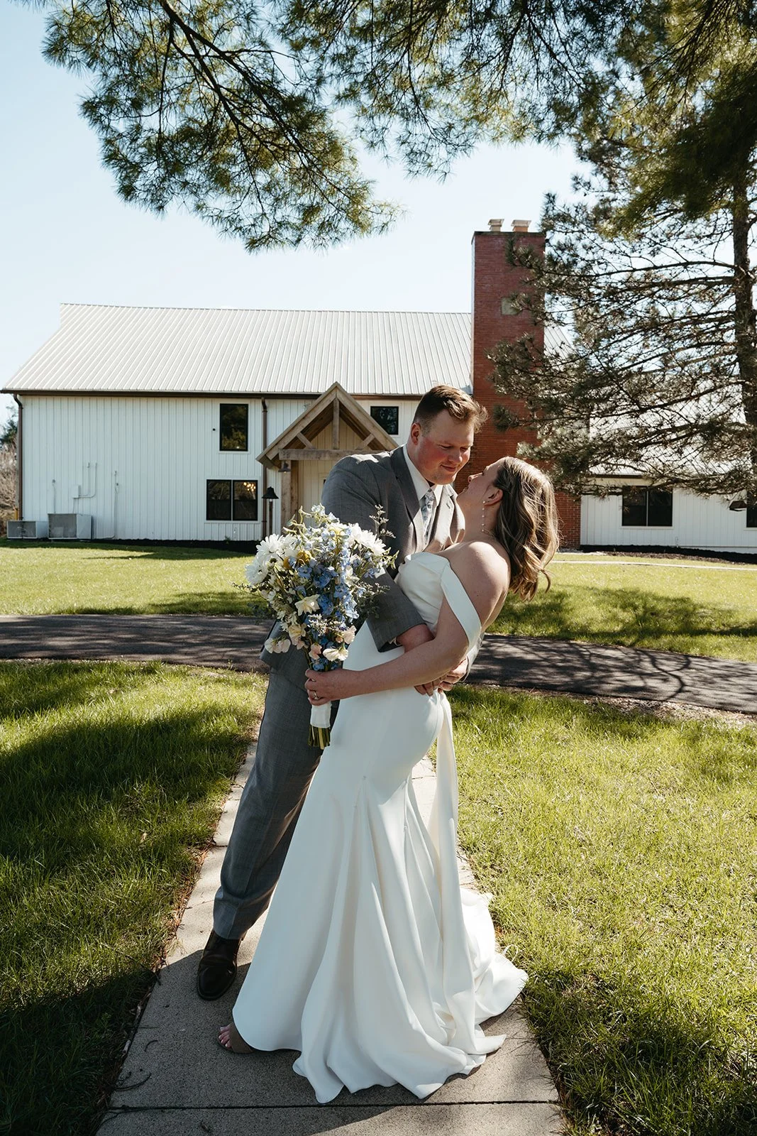 A newlywed couple in wedding attire sharing an embrace outdoors on a sunny day, with a white house and green lawn in the background.