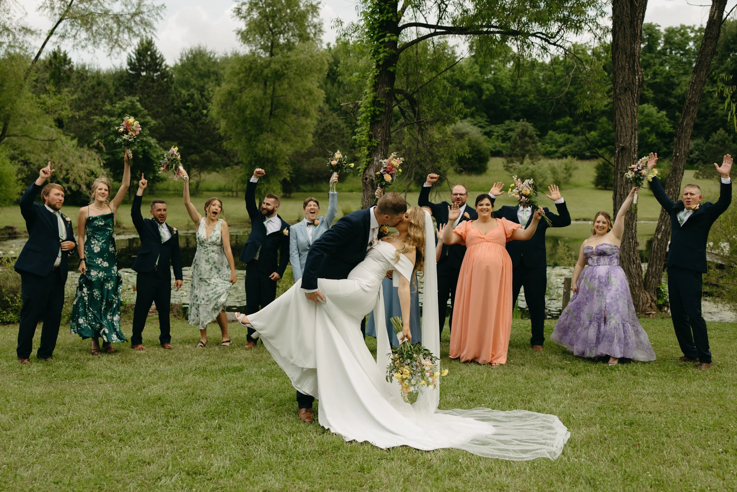 A newlywed couple kissing while standing on a grassy area surrounded by friends and family, celebrating their wedding outdoors with trees and a pond in the background.