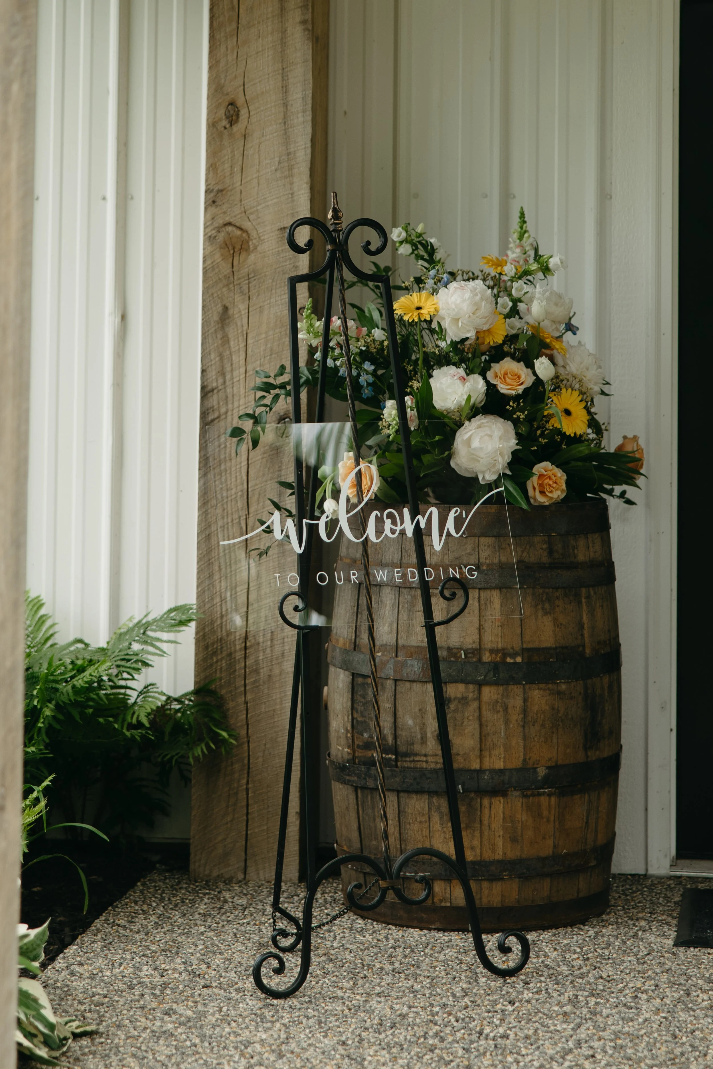 Welcome sign and flower arrangement at a wedding entrance, featuring a glass sign with white script and a wooden barrel with colorful flowers.